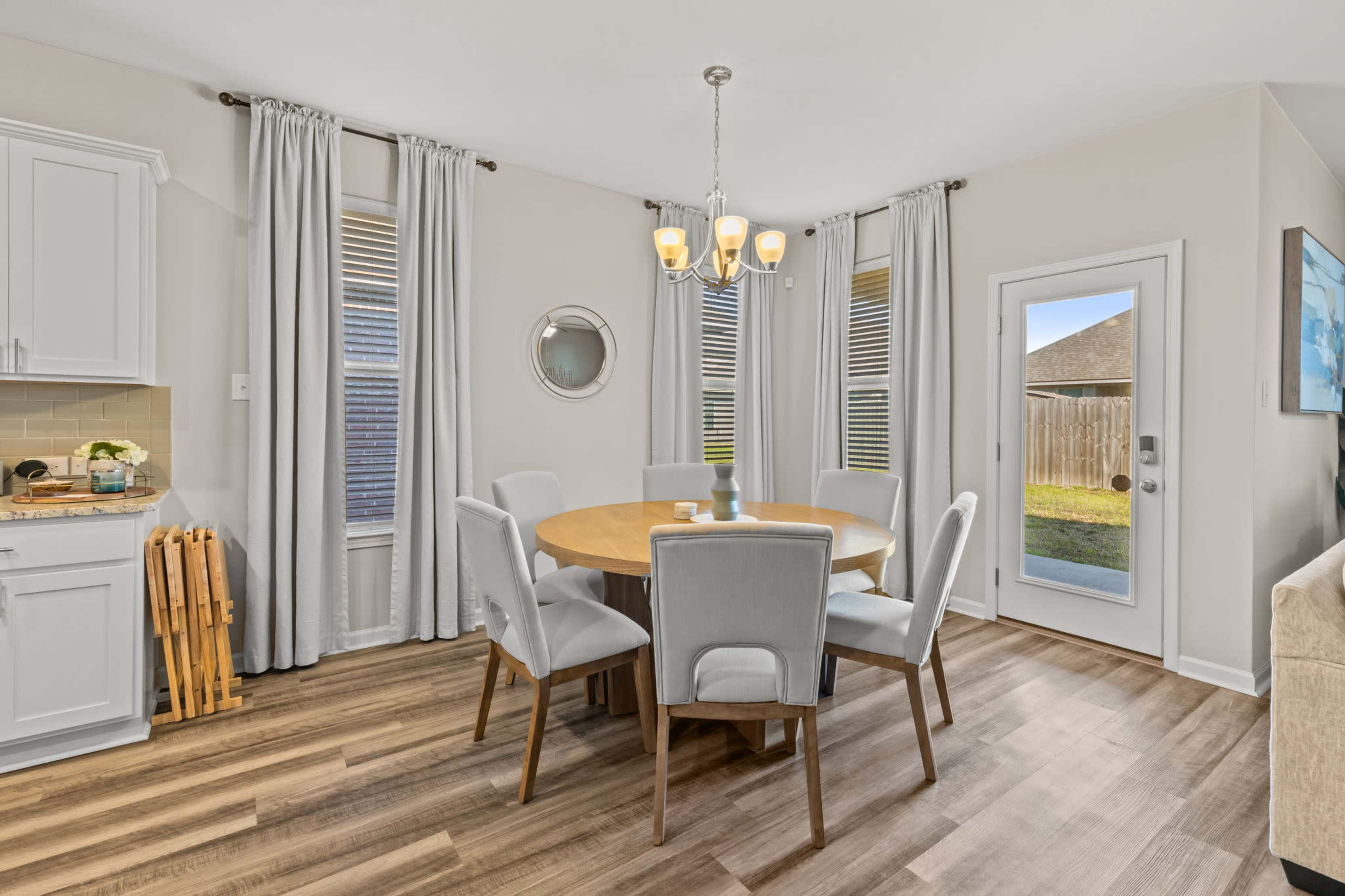 A modern dining area features a round wooden table surrounded by six upholstered chairs, with large windows and a sliding glass door that opens to a backyard.