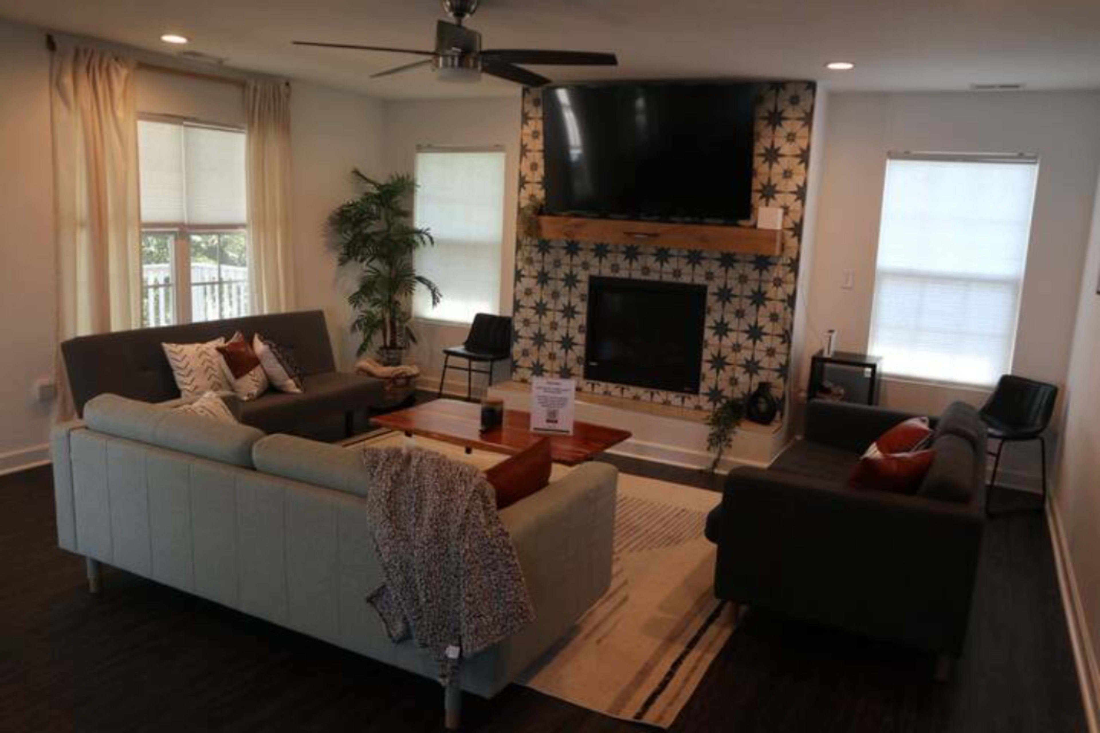 A modern living room features a central coffee table surrounded by two sofas, a television mounted above a patterned fireplace, and large windows allowing natural light.