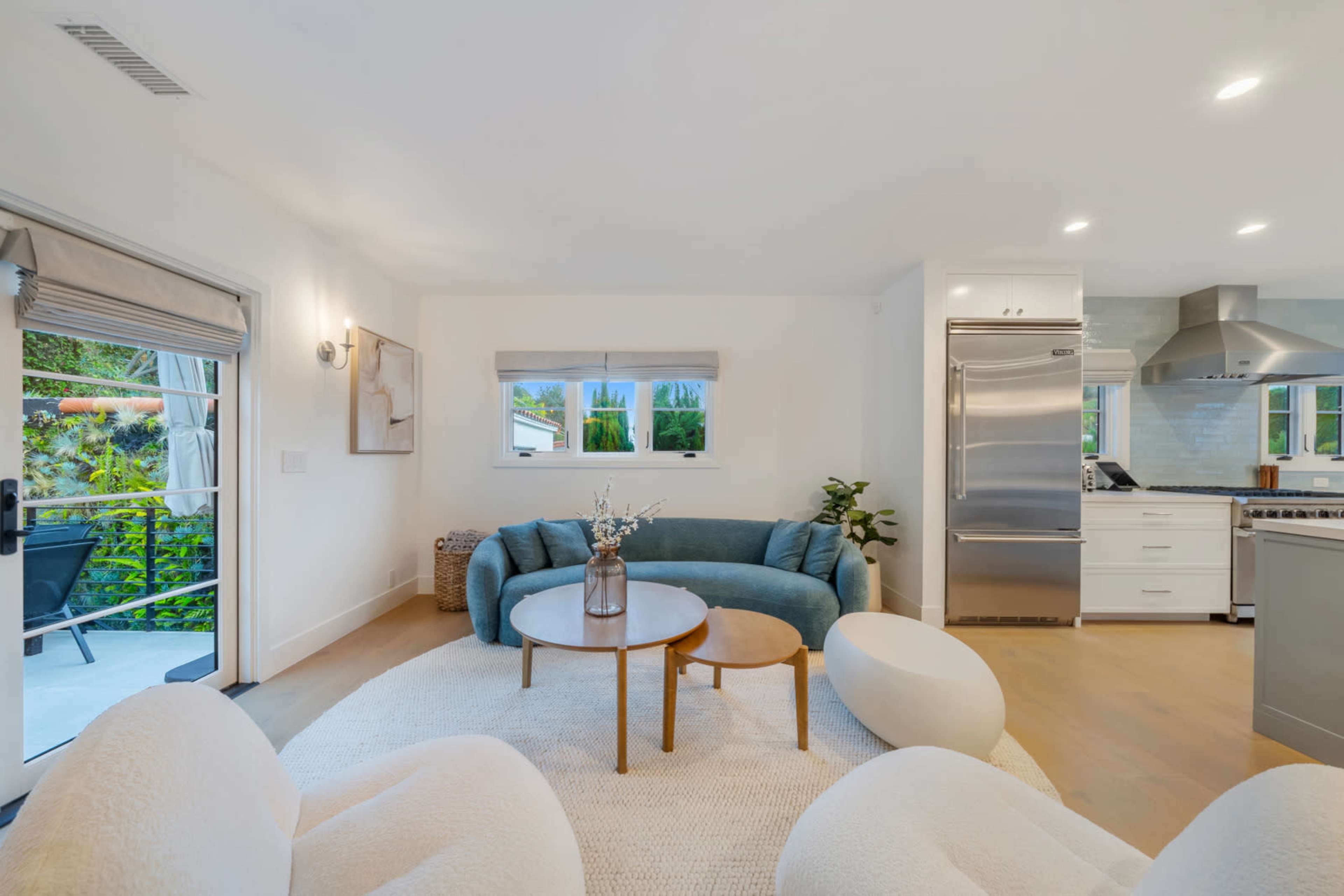 The image shows a modern living room with a blue sofa, two round tables, and white chairs, featuring large windows that overlook a garden.