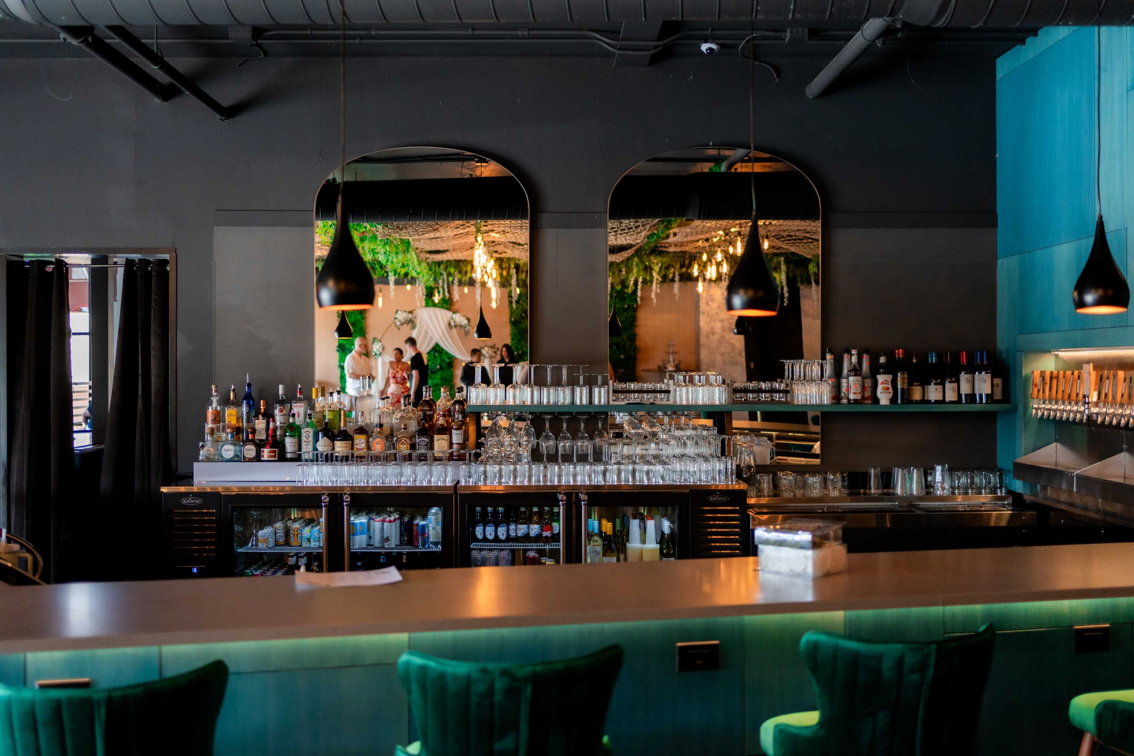 The image depicts a bar area with a sleek countertop, an array of liquor bottles behind the counter, and decorative mirrors reflecting the ambiance.