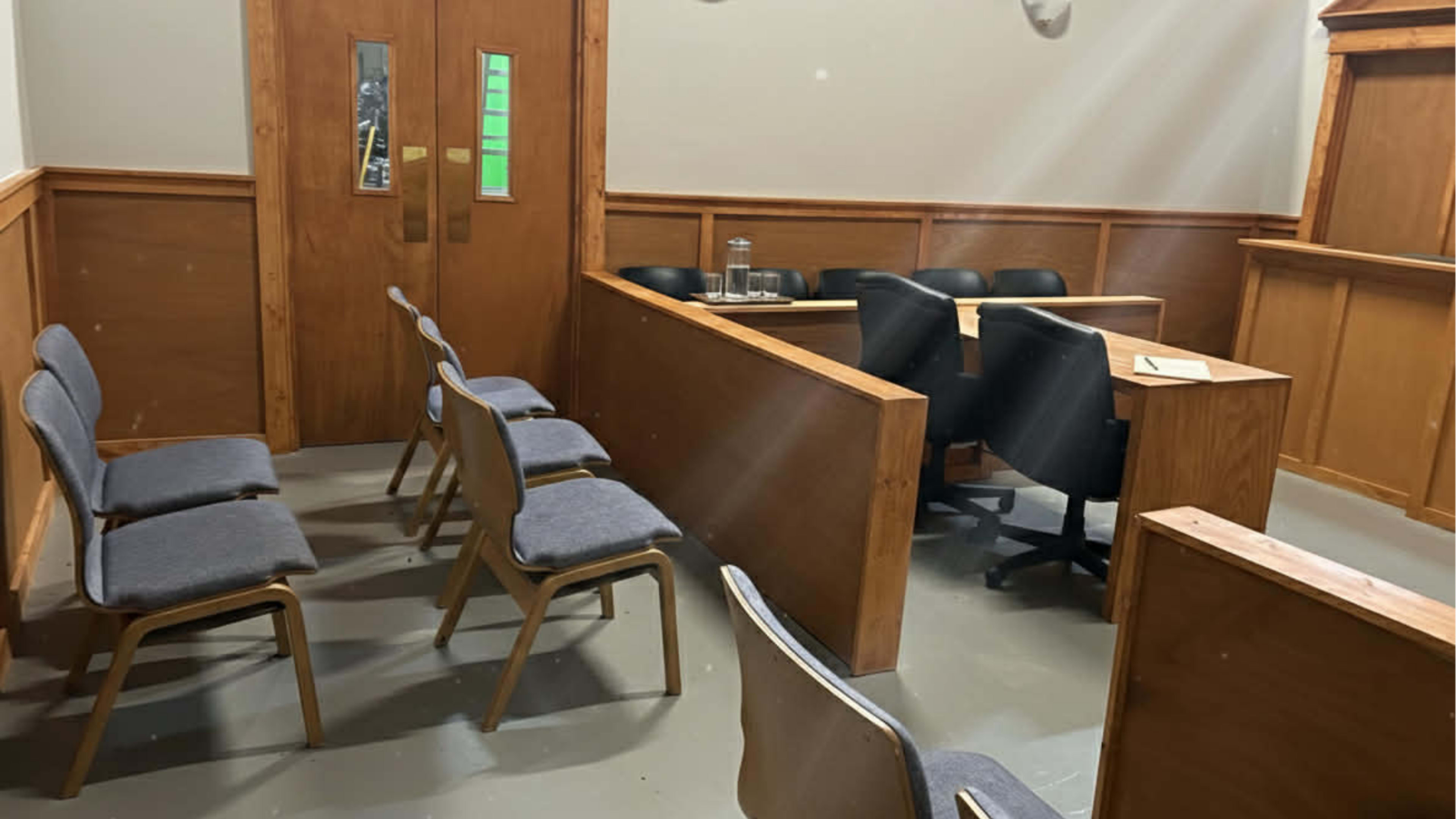 The image shows a small, empty courtroom with wooden paneling, a judge's bench, a table for legal counsel, and adjacent seating for the public.