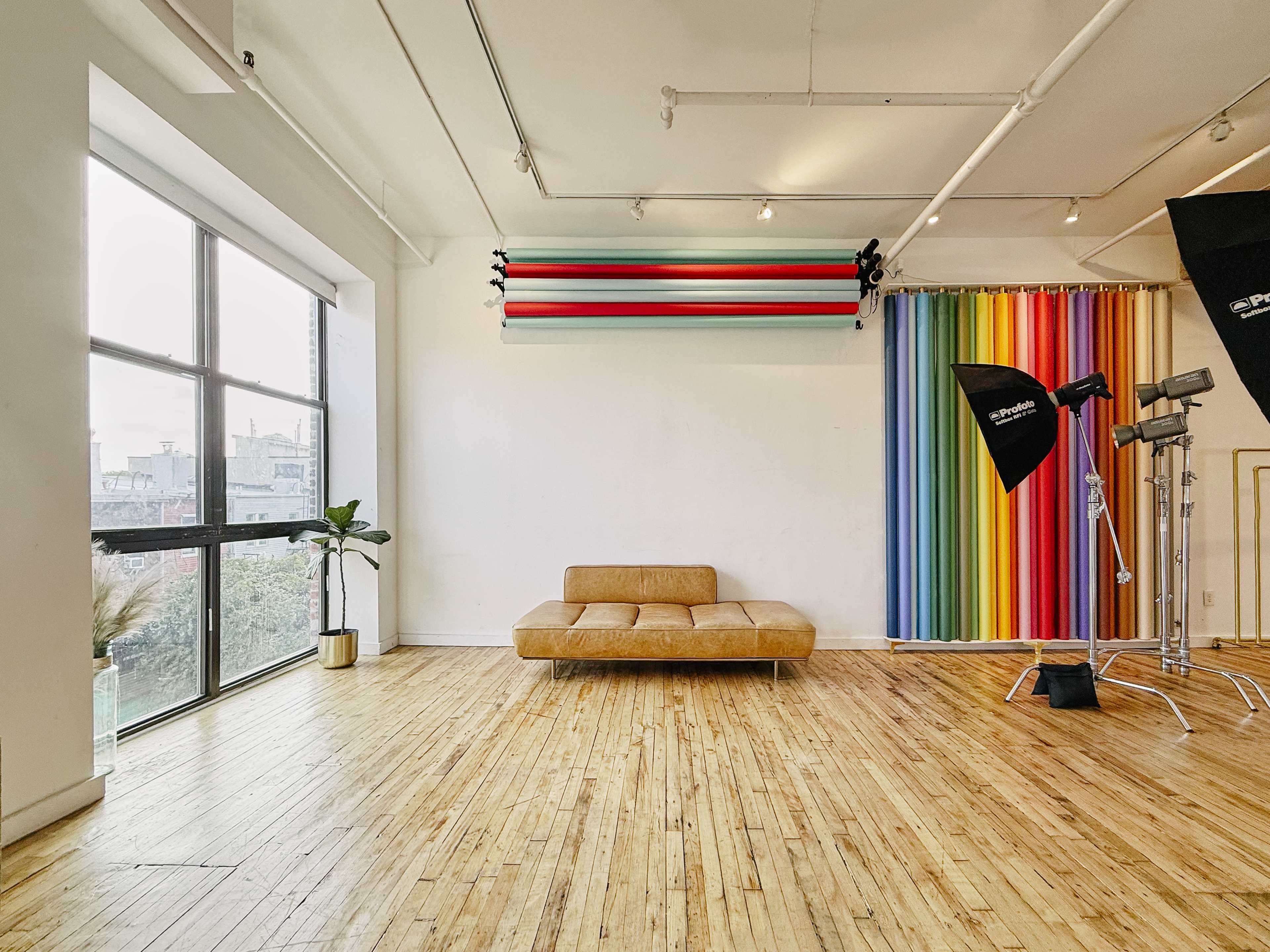 The image shows a photography studio with wooden flooring, a large window, a beige sofa, and a colorful backdrop with stripes.