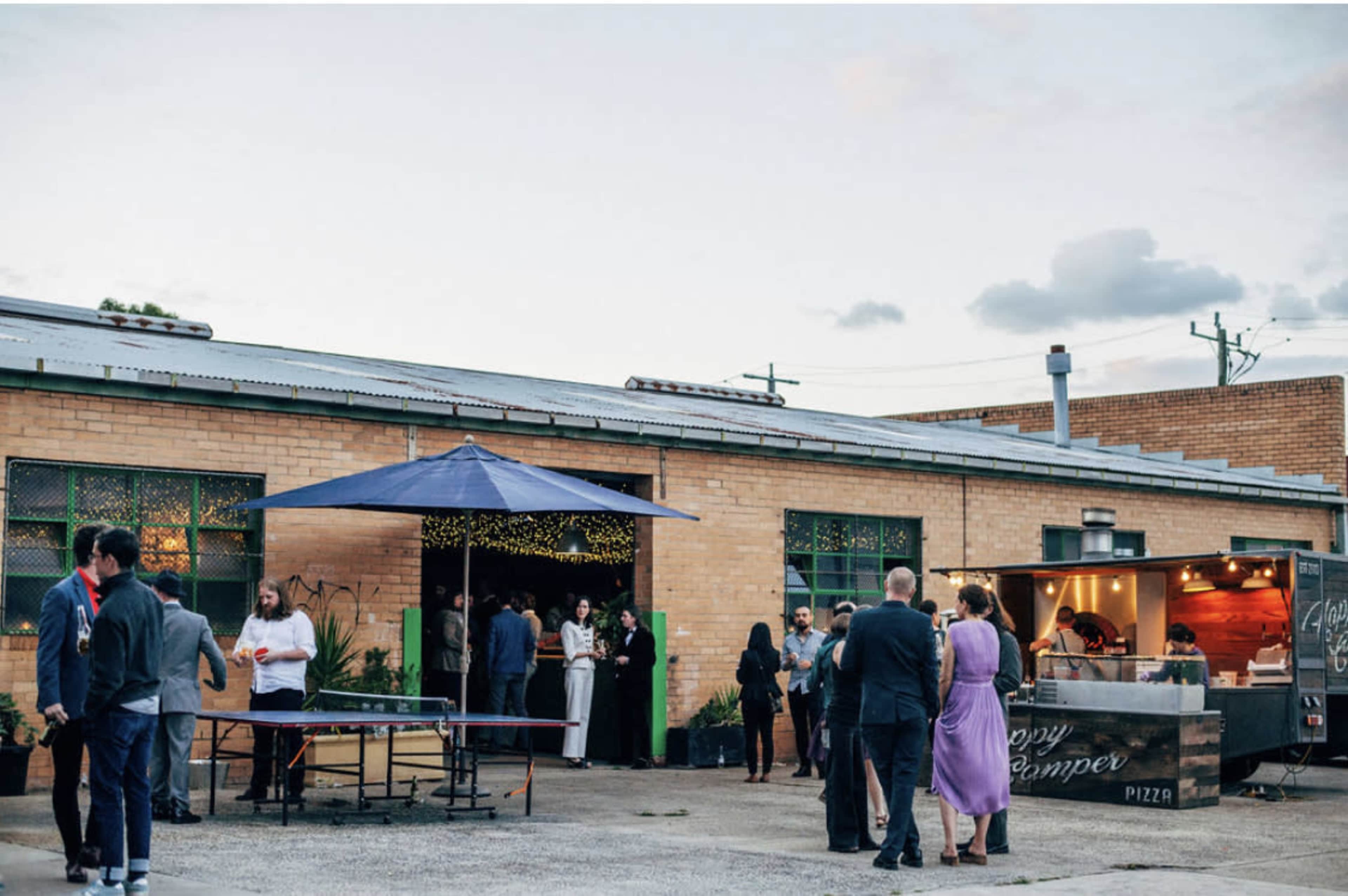 A group of people gathers outside a brick building with string lights and a food truck during an evening event.