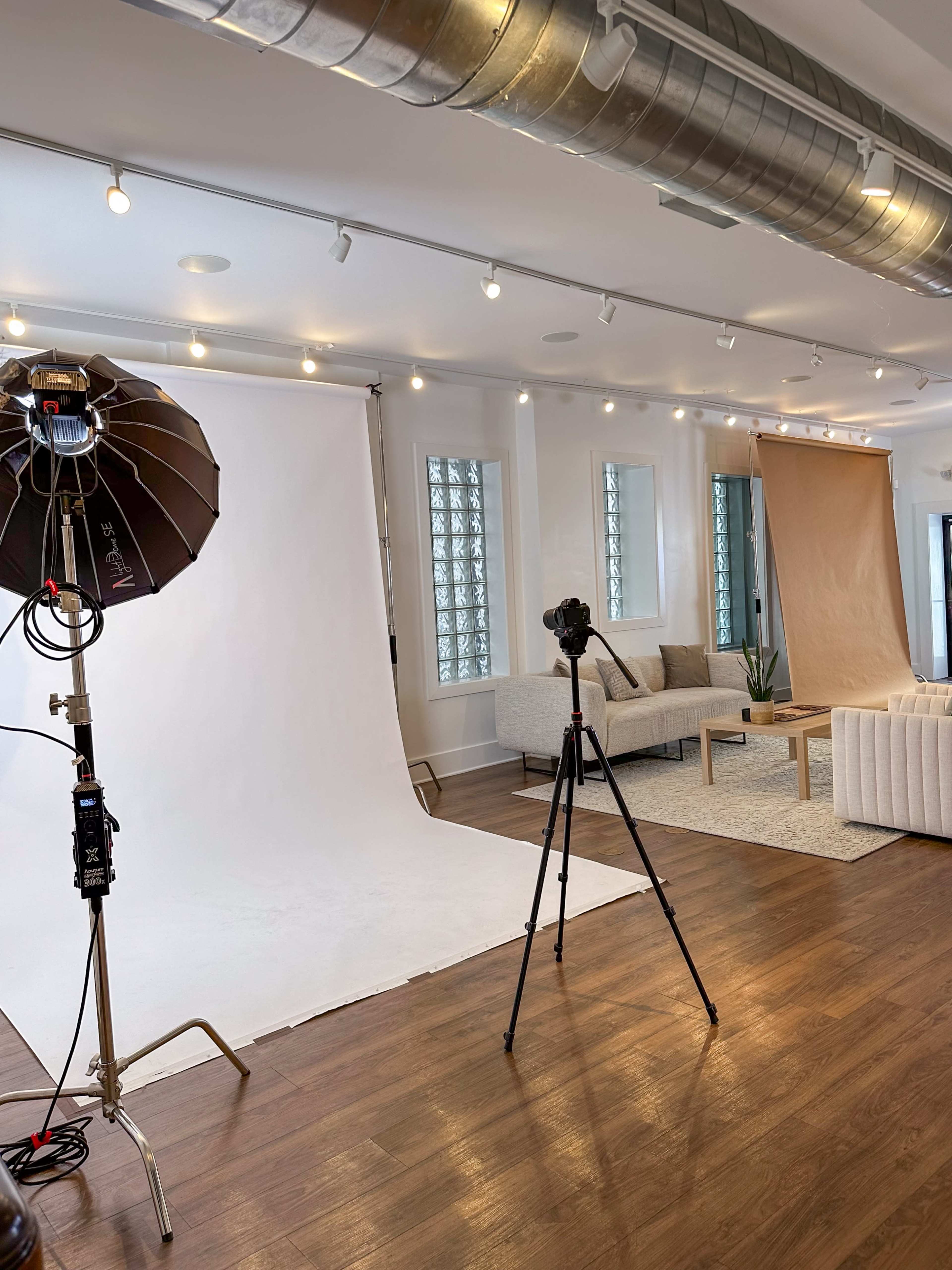 The image shows a photography studio with a white backdrop, a lighting setup, a camera on a tripod, and a seating area featuring a sofa and decorative elements.