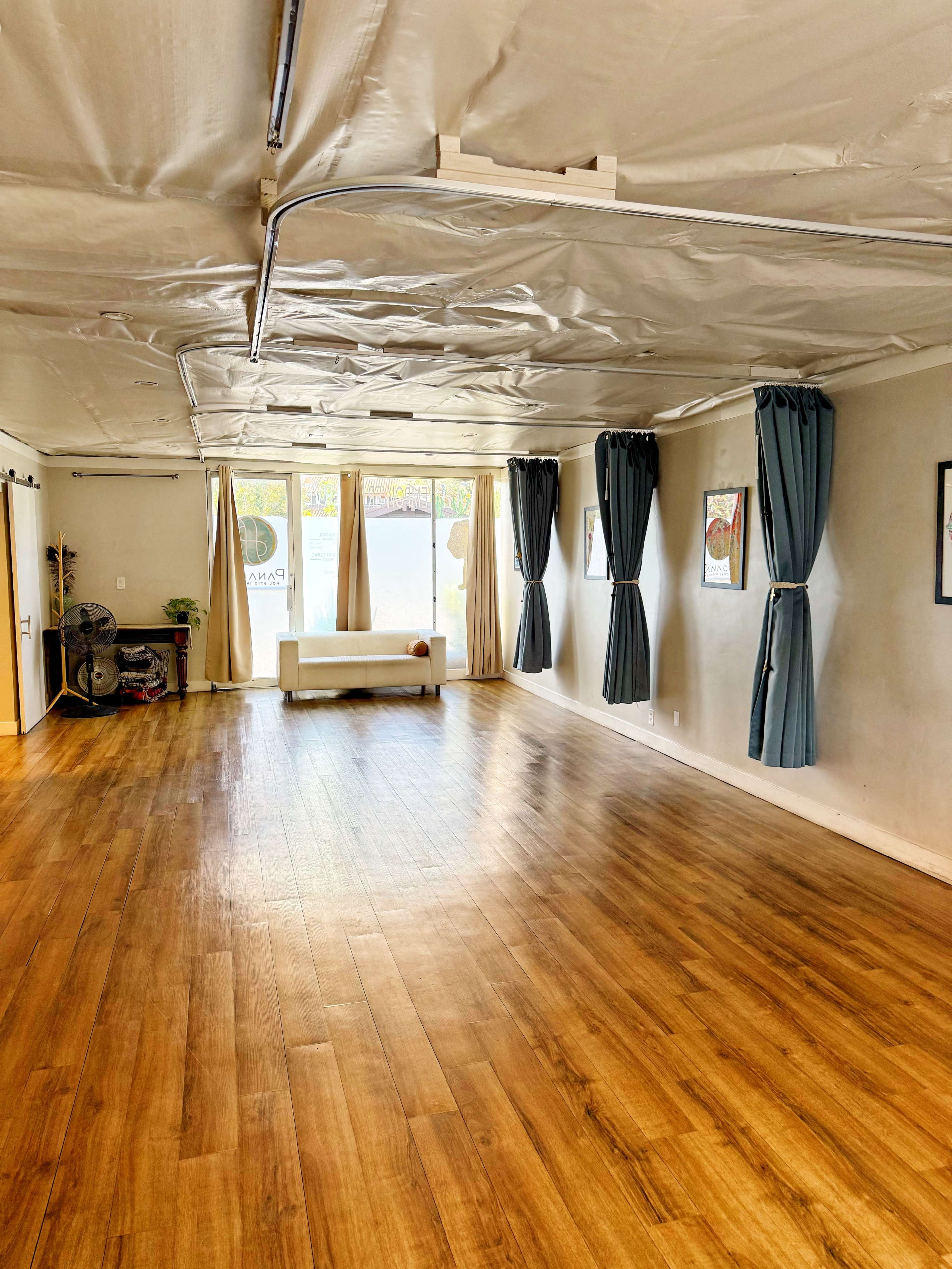 The image shows an empty room with wooden flooring, large windows, and blue curtains, featuring a white sofa and several framed pictures on the walls.