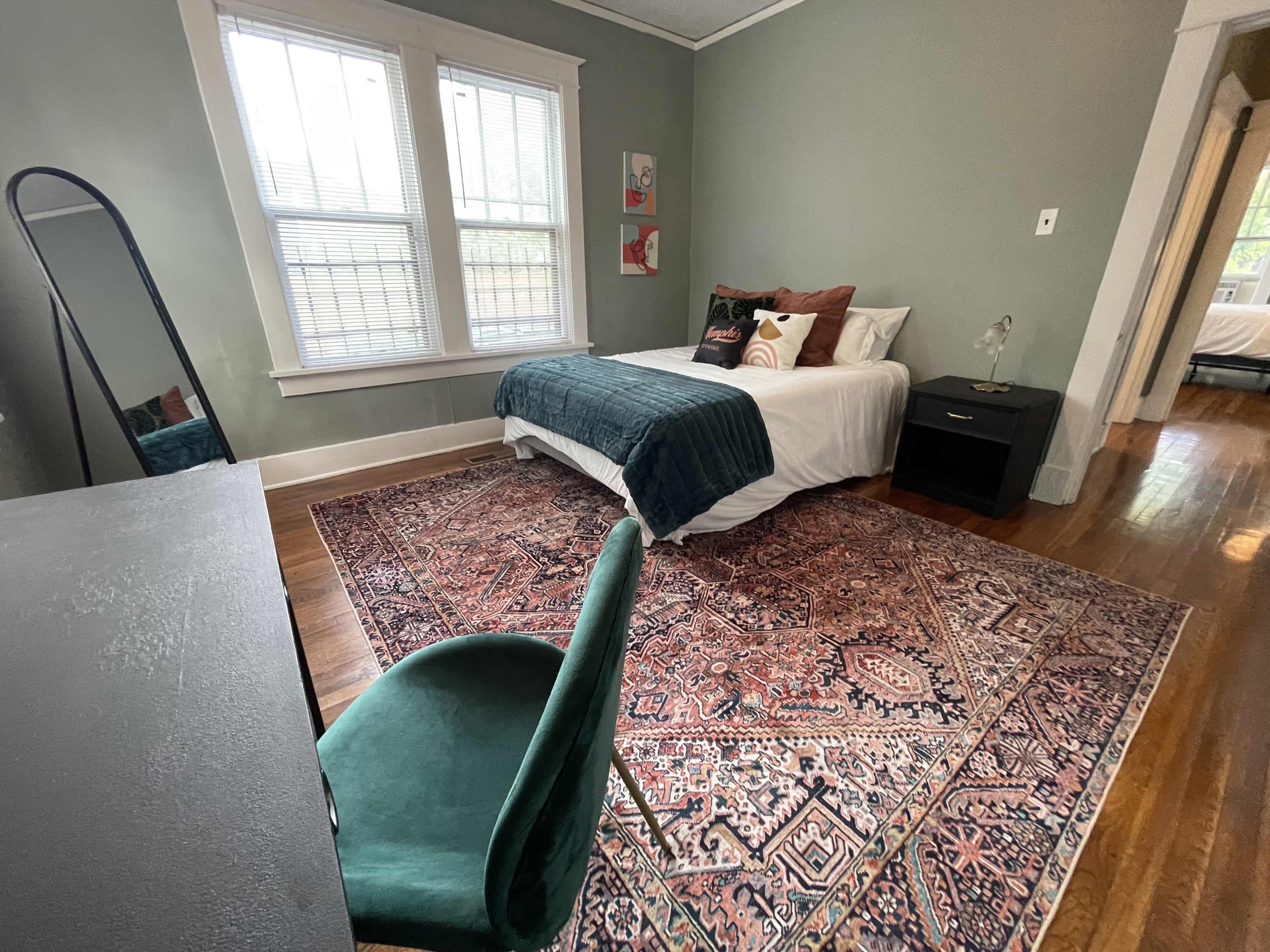 A cozy bedroom featuring a bed with a green throw blanket, a desk and chair, and a large area rug on polished wooden floors.