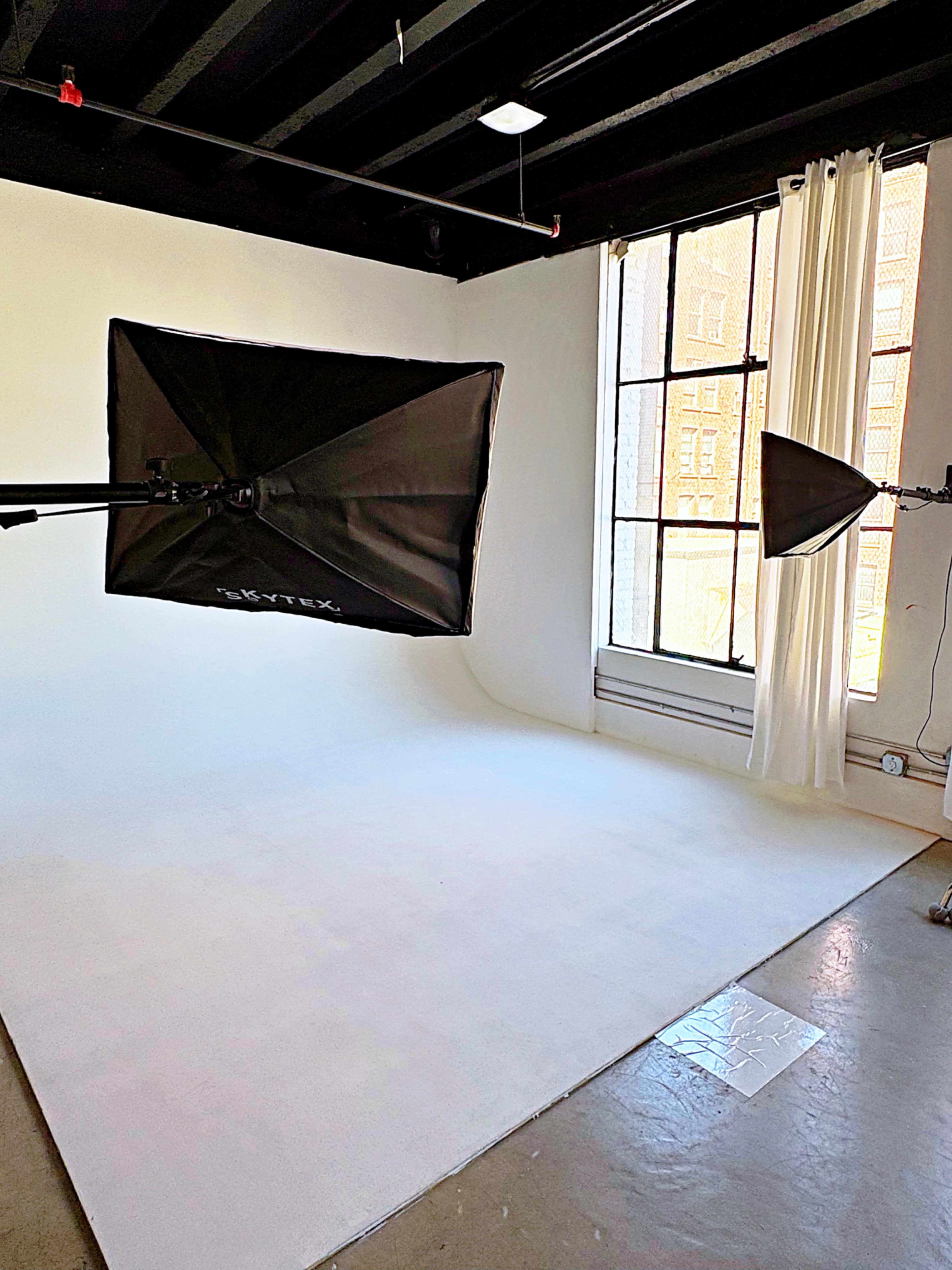 The image shows a photography studio with a white backdrop, softbox lights mounted on adjustable arms, and large windows covered by sheer curtains.