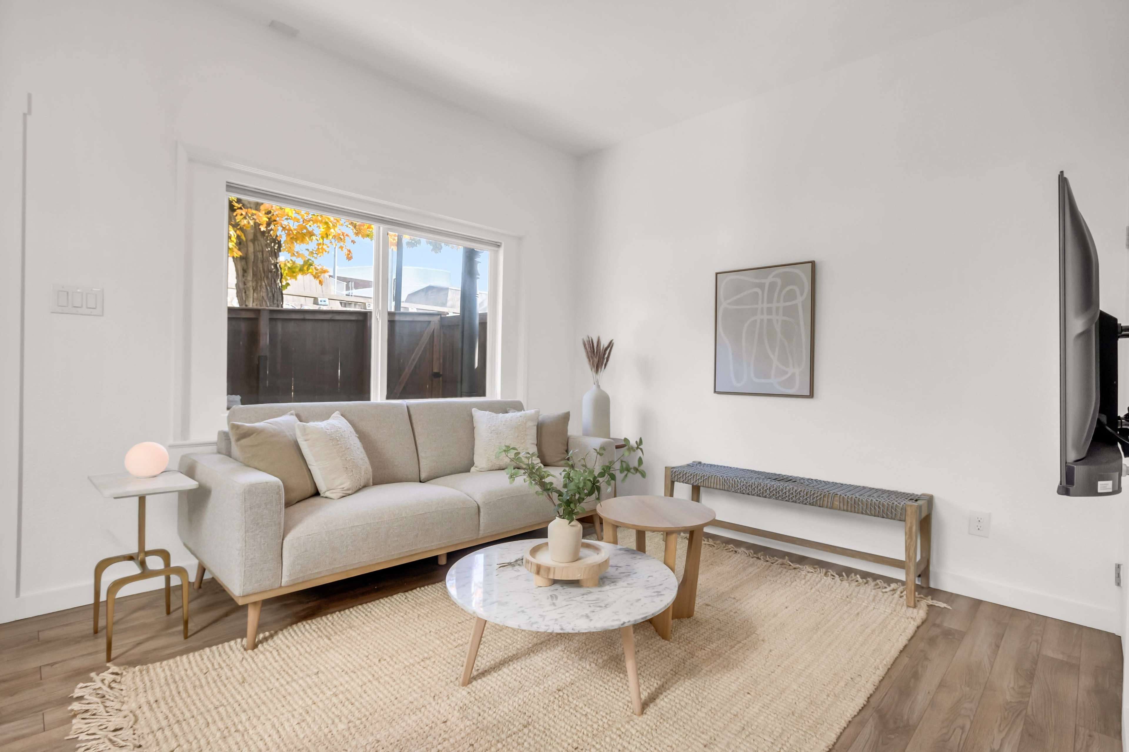 A light-filled living room features a beige sofa, a round marble coffee table, a woven rug, and a minimalist art piece on the wall.