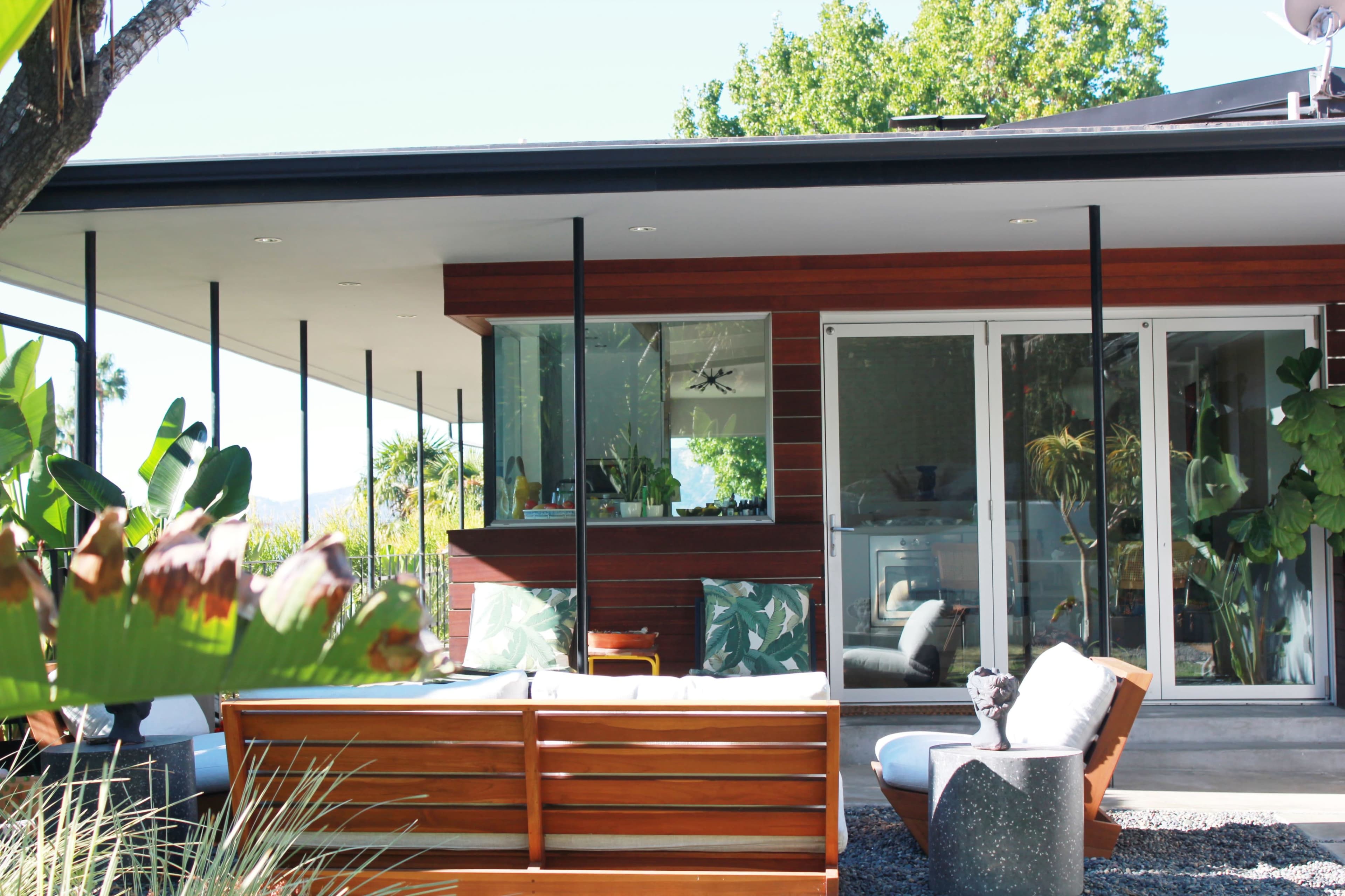 A modern house features large glass windows and a wooden patio surrounded by greenery.