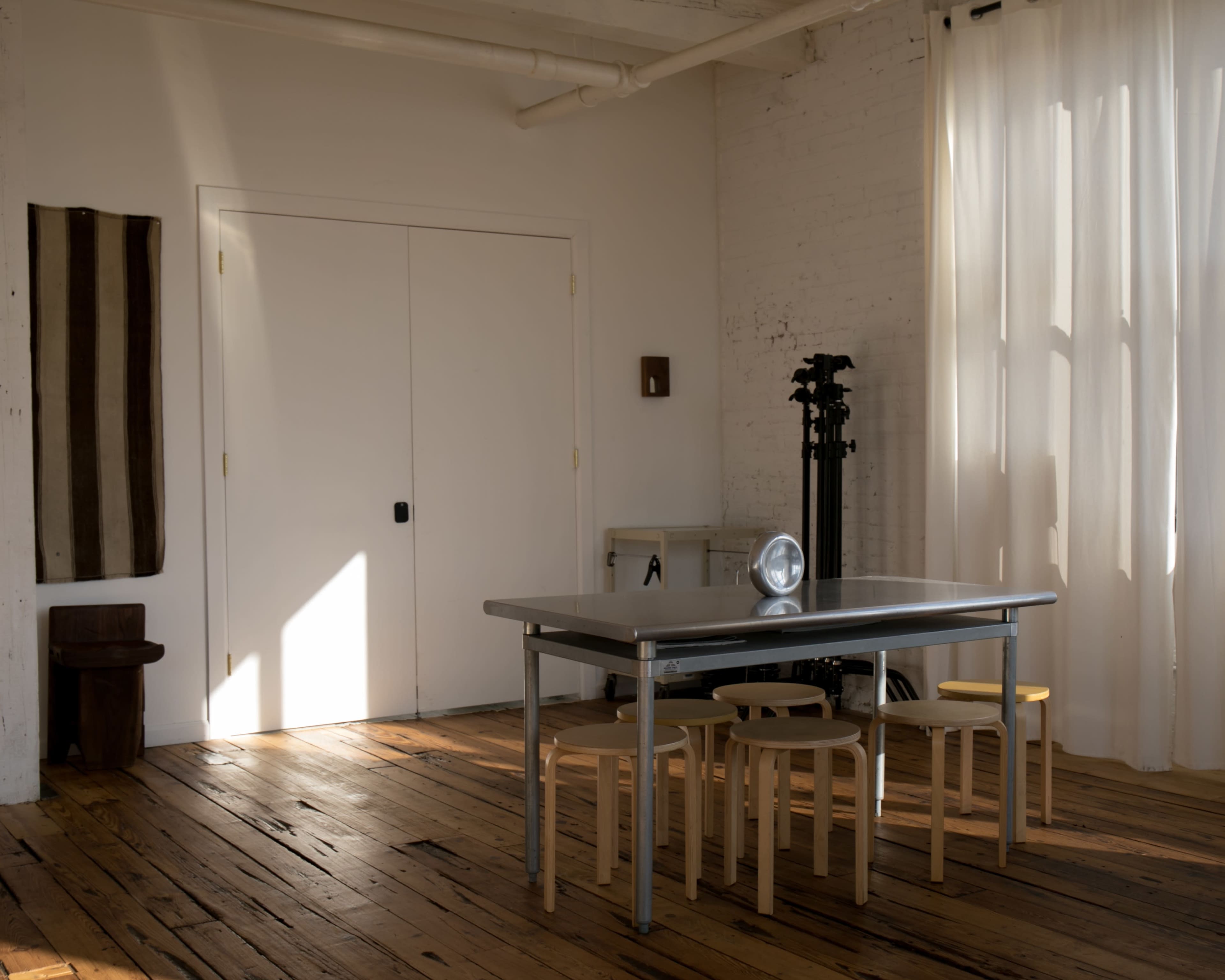 A minimalist interior features a metallic table surrounded by four wooden chairs in a sunlit room with white walls and a large window.