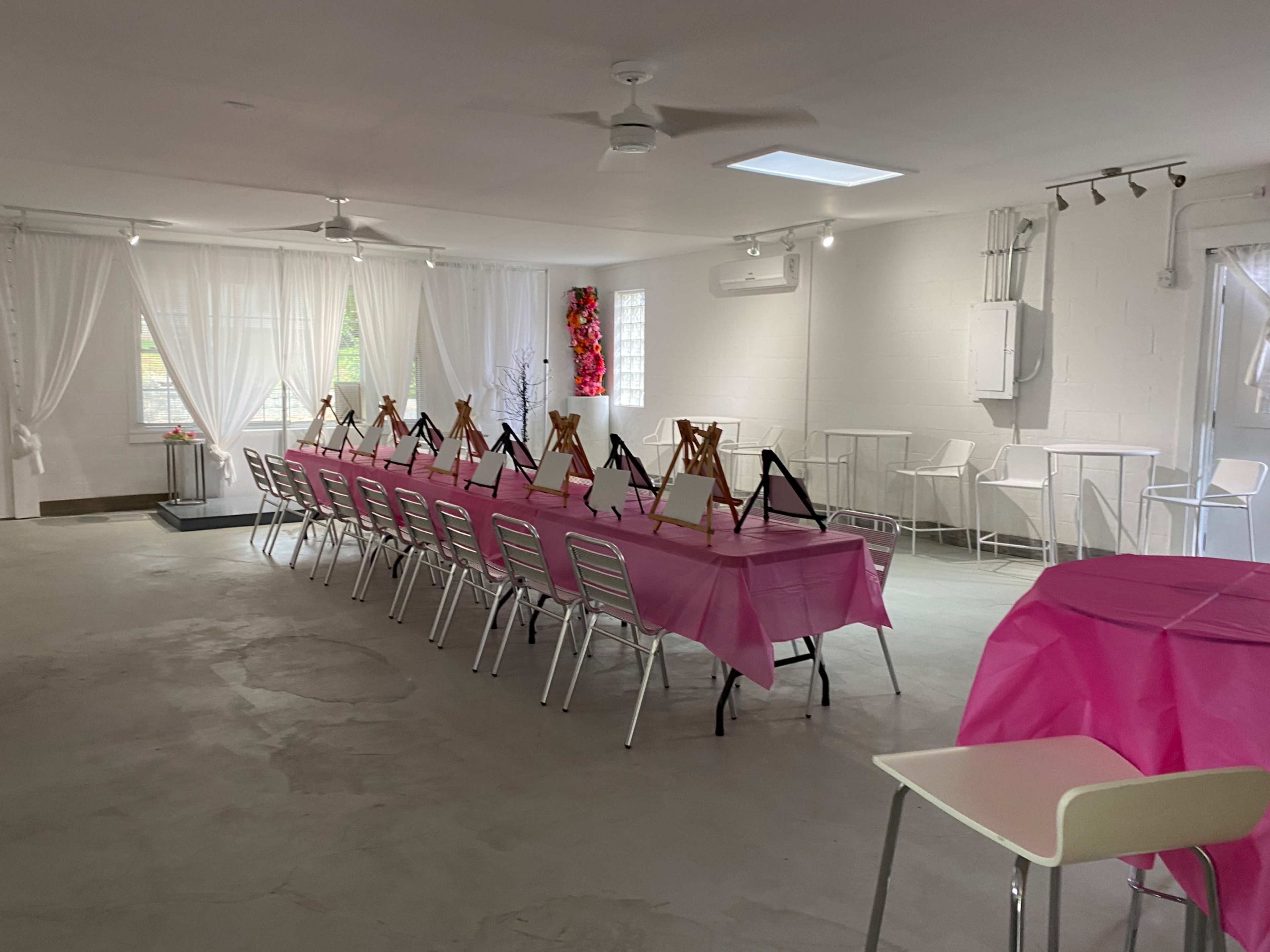 A spacious room is set up with a long table covered in pink cloth, surrounded by metal chairs and decorated easels.