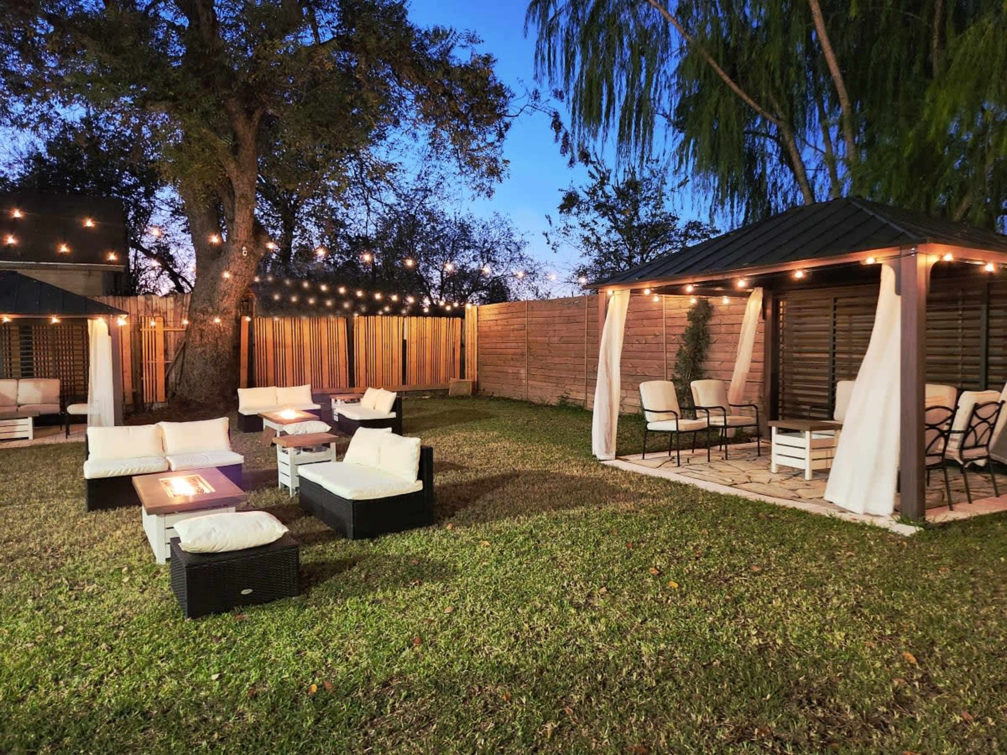 A backyard seating area with illuminated string lights, a gazebo, and lounge furniture arranged on grass.