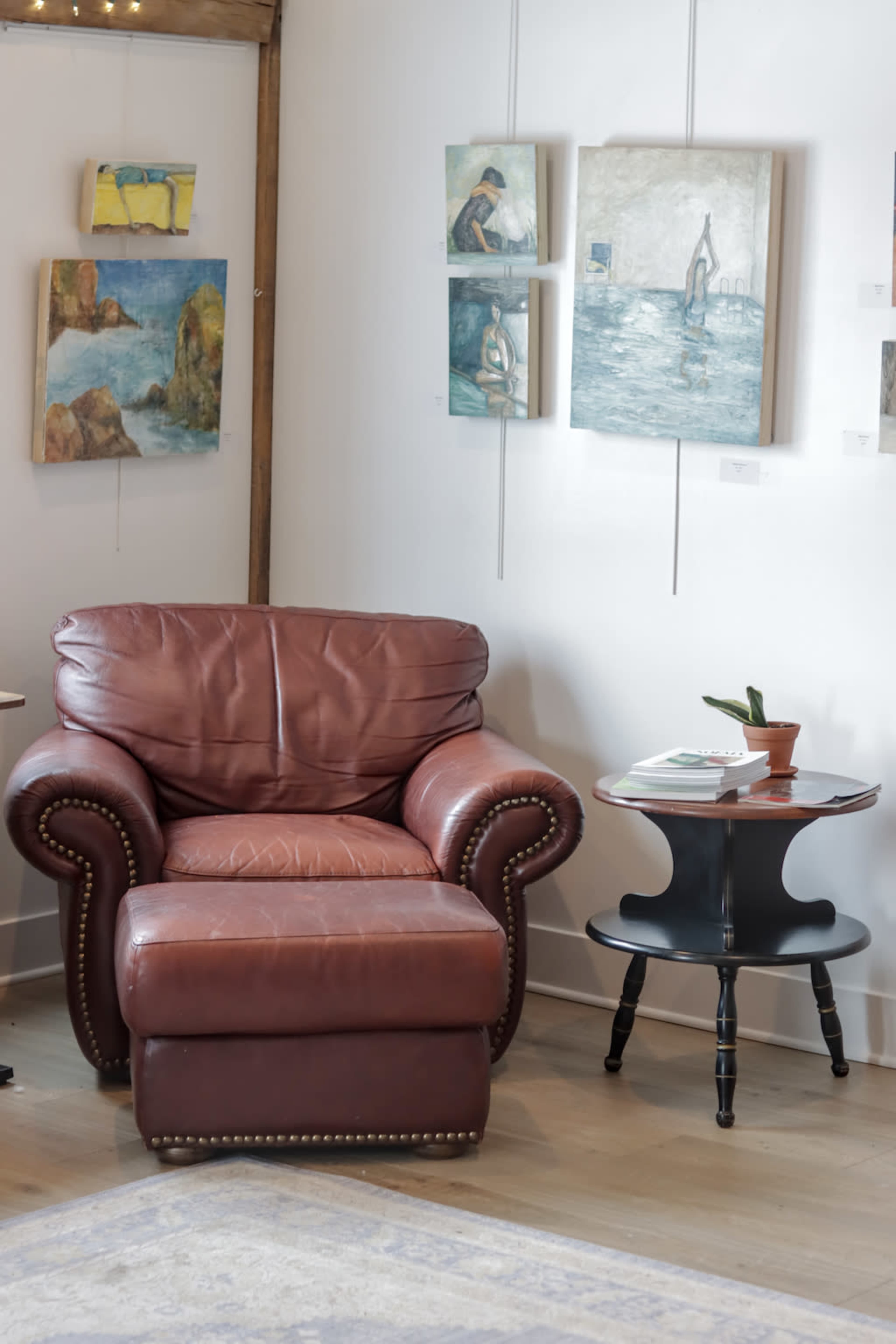 The image shows a brown leather armchair next to a round black side table with a small plant and books in an art gallery with paintings on the walls.