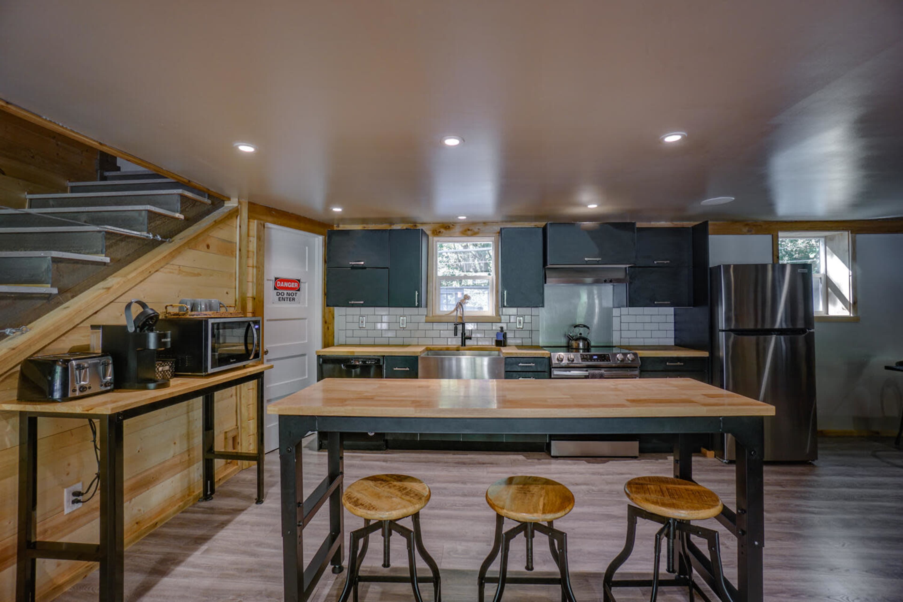The image shows a modern kitchen with wood and metal accents, featuring a central island with stools, stainless steel appliances, and wooden cabinetry.