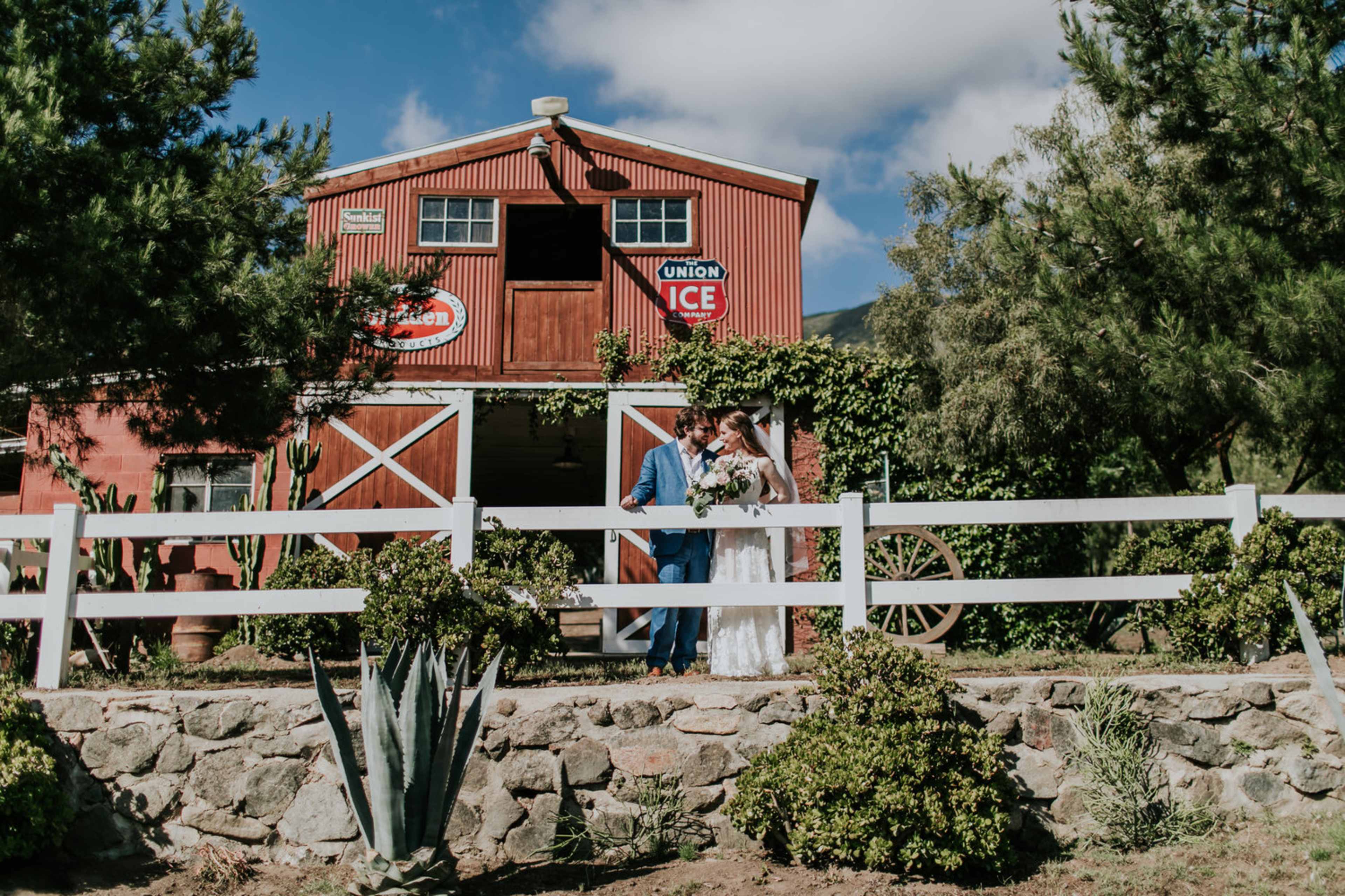A couple stands together in front of a red barn adorned with signs, surrounded by greenery and decorative plants.