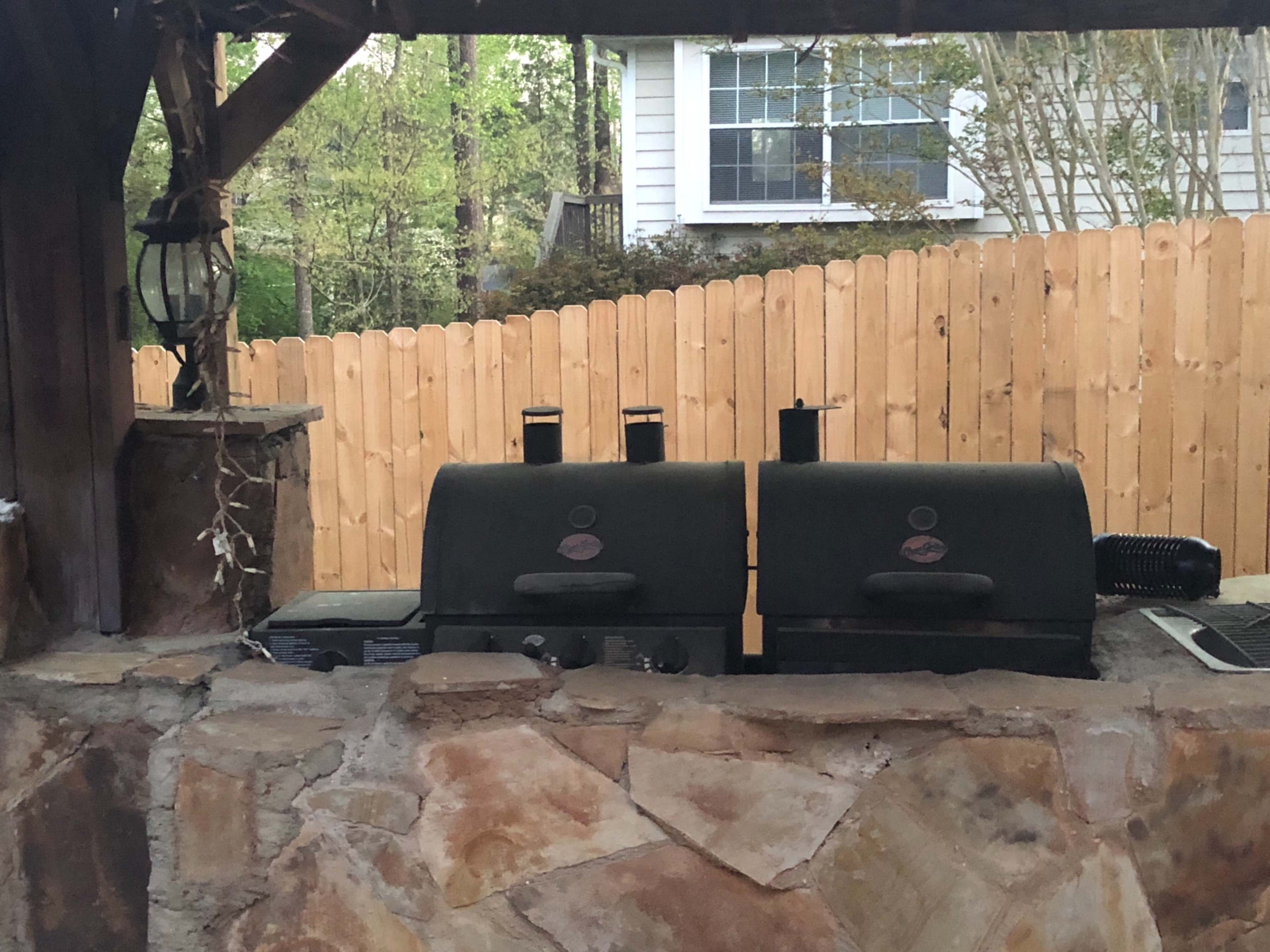 A stone outdoor grilling area featuring two green grills under a wooden gazebo.