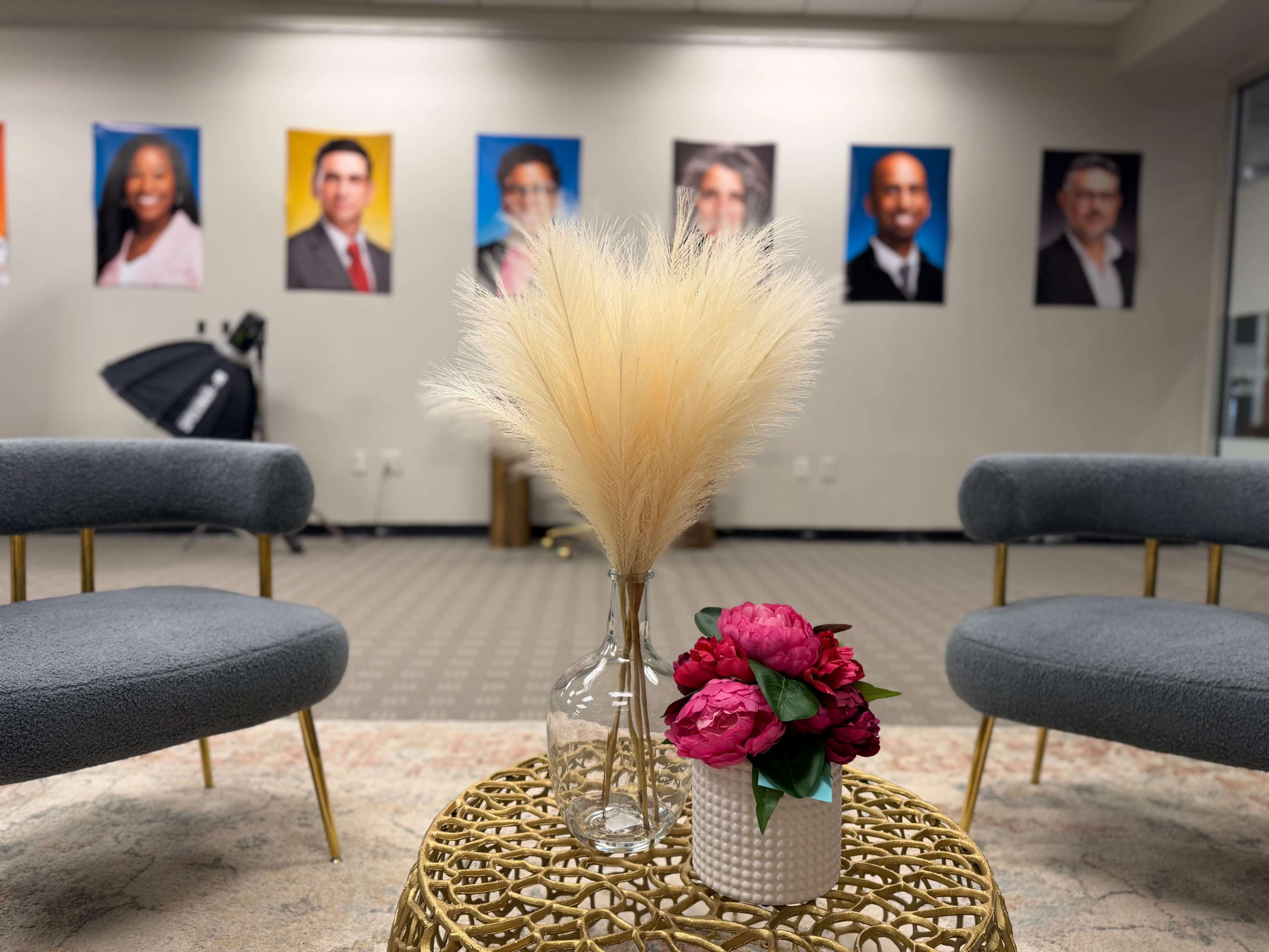 The image shows a modern office setting featuring two blue chairs facing a round table with decorative flowers and a large arrangement of dried plants, while portraits of individuals are displayed on the wall behind.