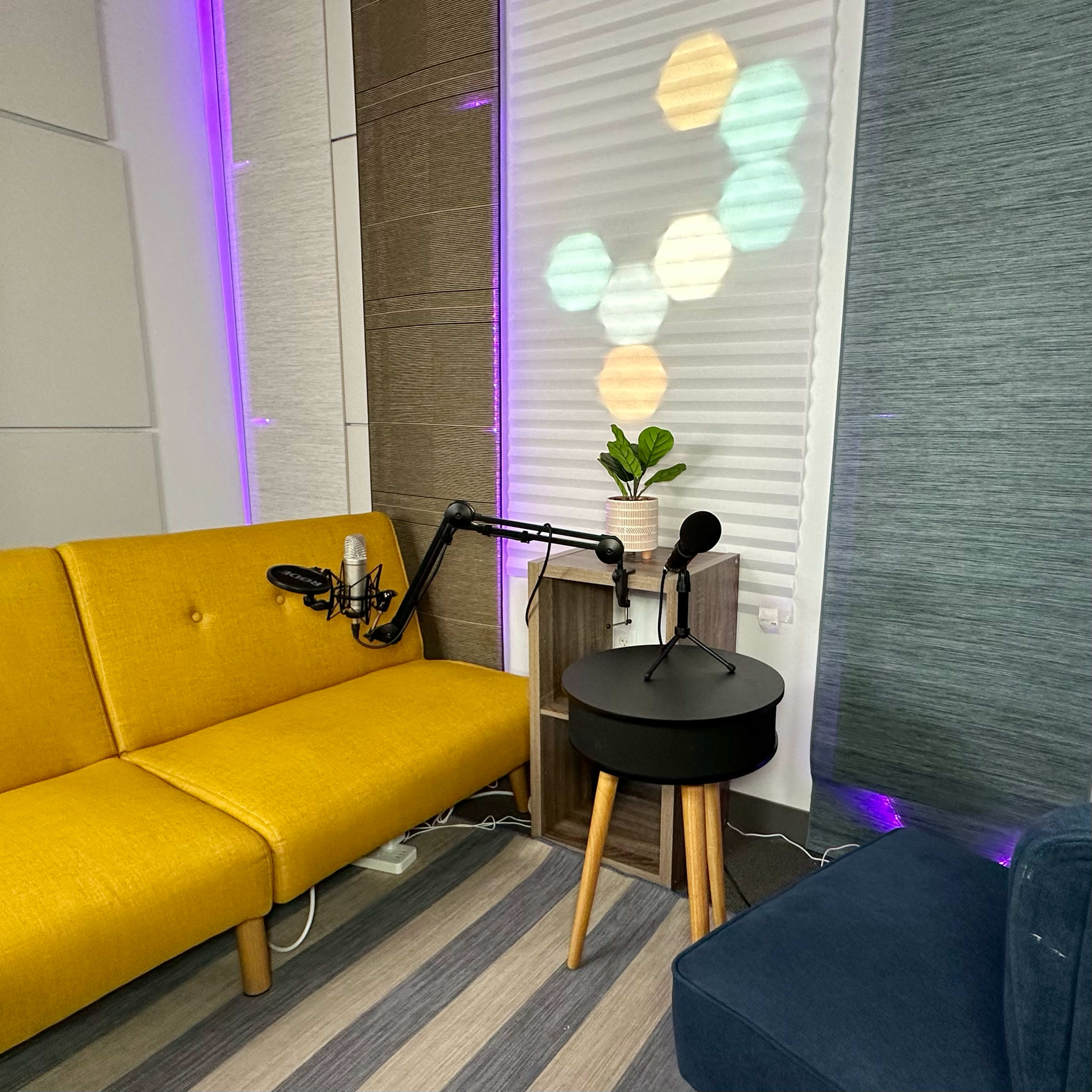 A cozy room features a yellow sofa, a dark round table, a microphone setup, and a small potted plant with colorful light reflections on the walls.