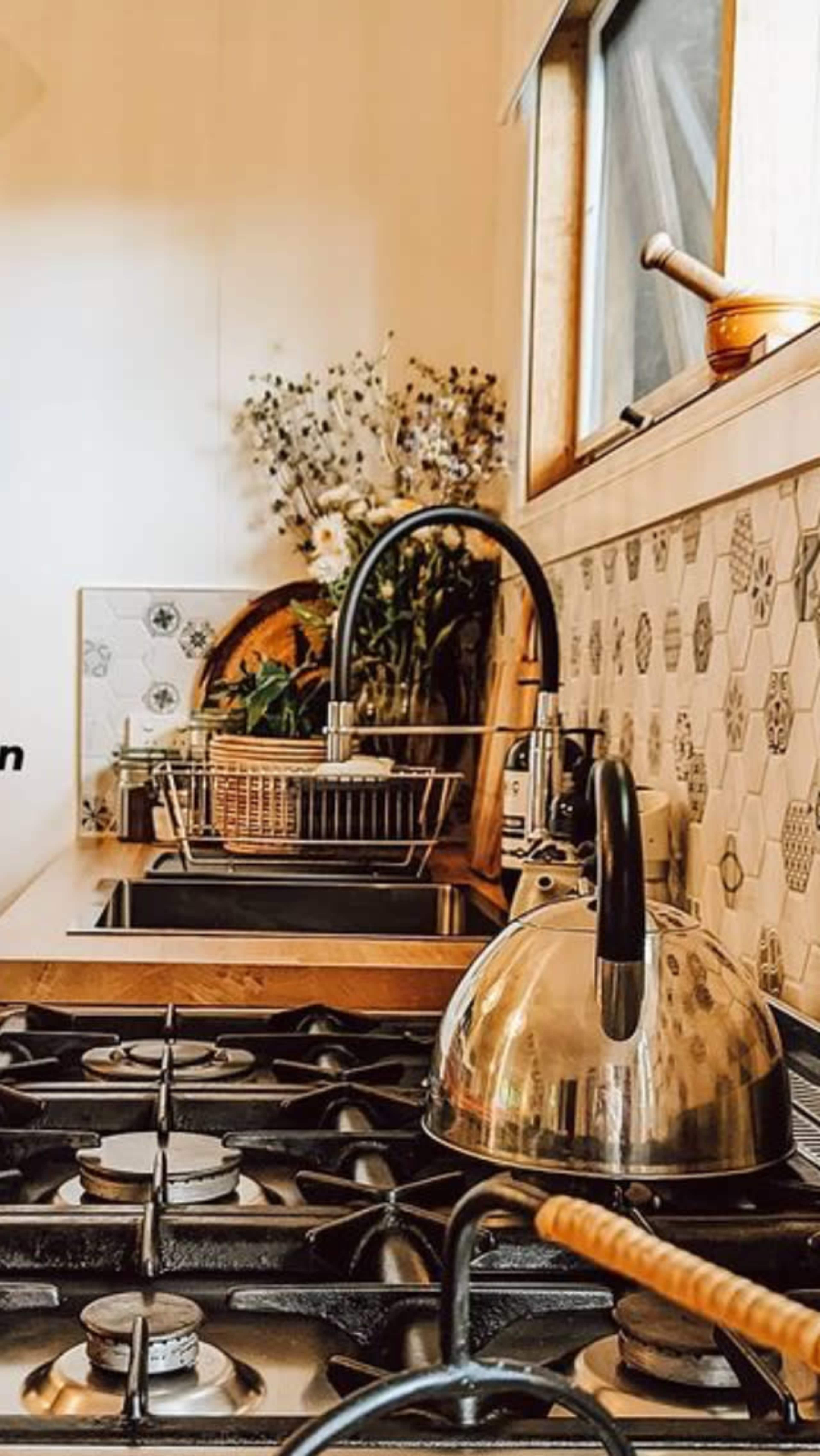 The image shows a kitchen countertop with a gas stove, a stainless steel kettle, a drying rack, and a vase of flowers in the background.