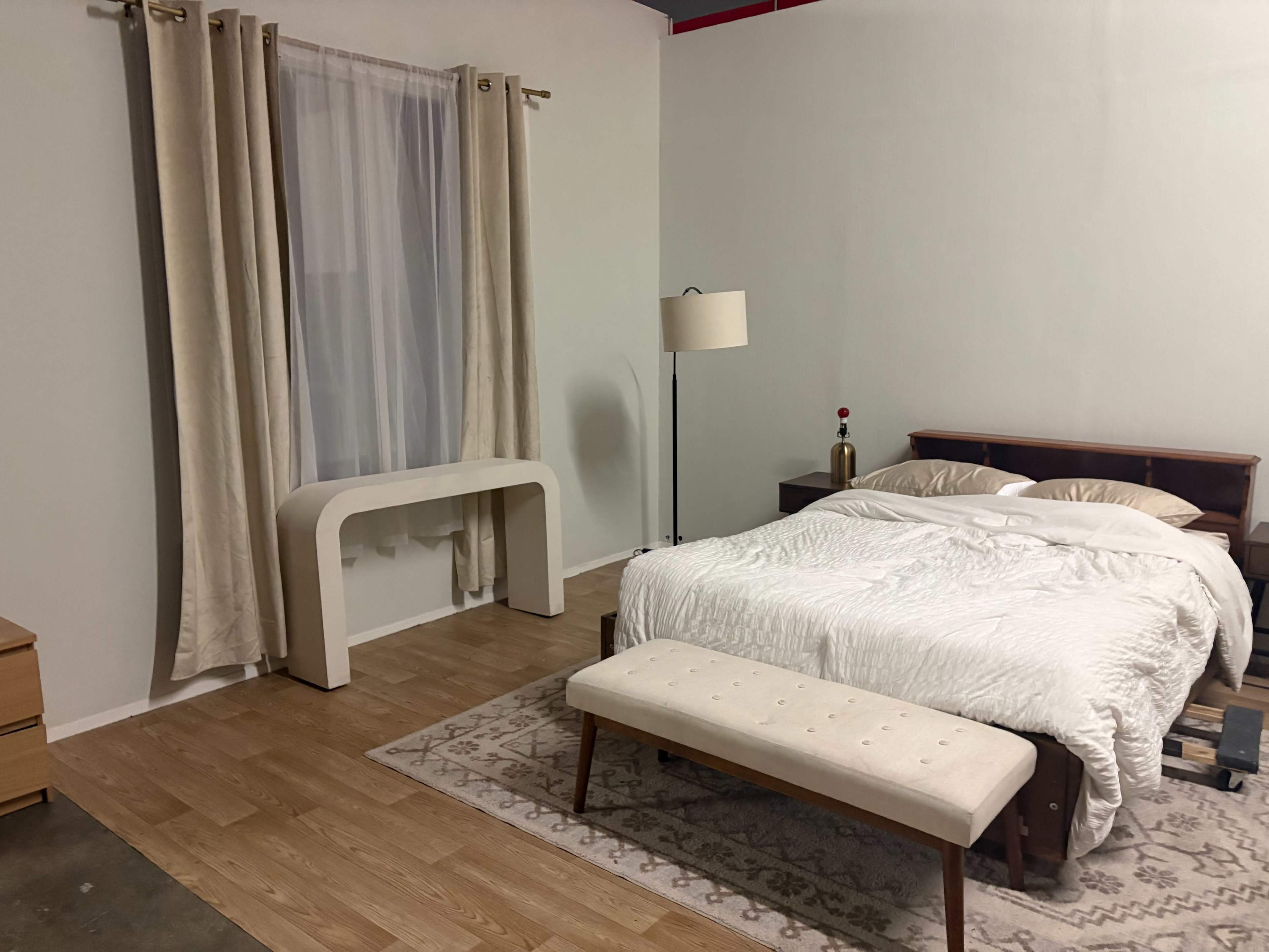 The image shows a minimally furnished bedroom with a bed, a bench at the foot, a side table, a lamp, and a desk against a wall, along with curtains over a window.