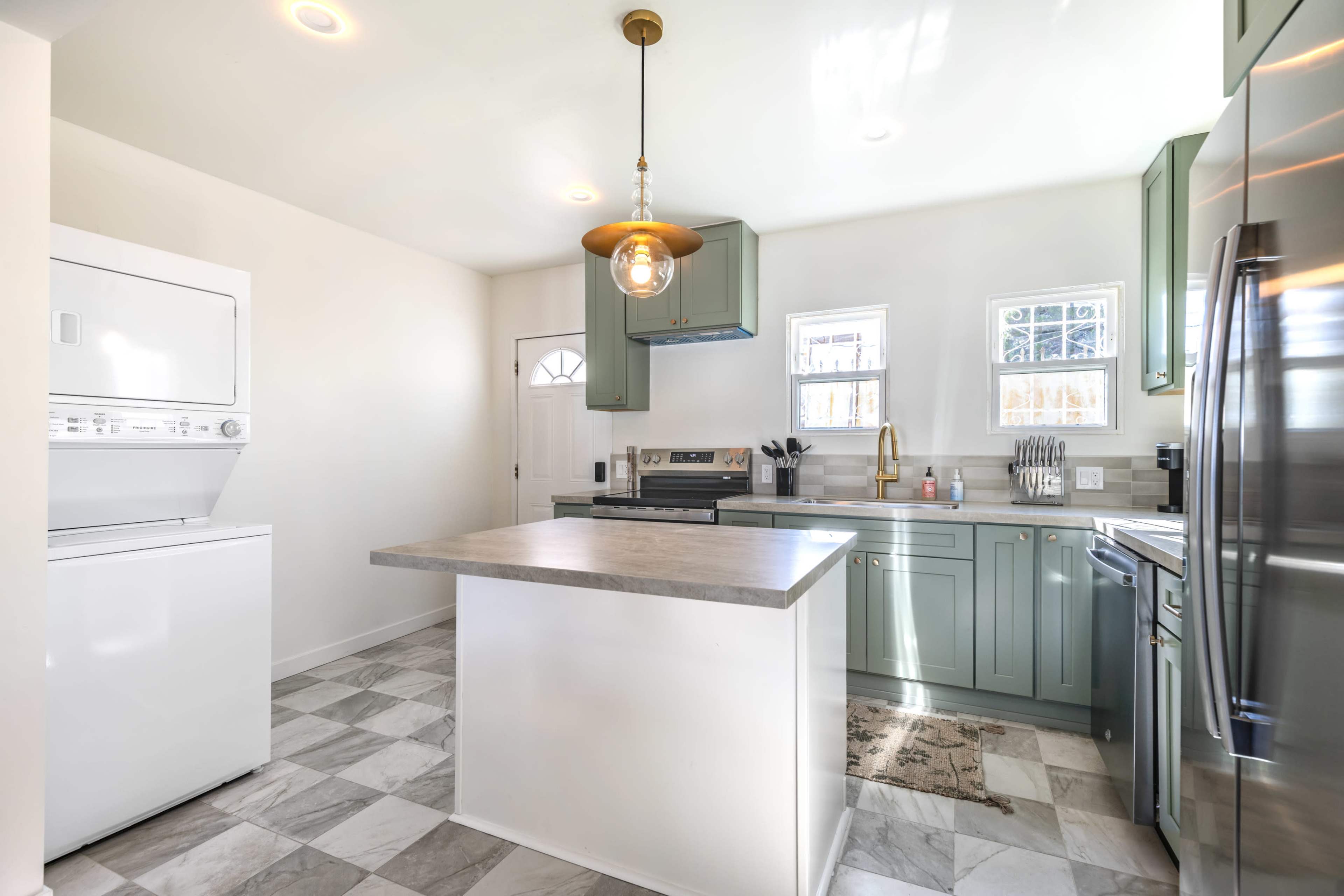 The image shows a modern kitchen featuring green cabinets, stainless steel appliances, a kitchen island, and a stacked washer and dryer.