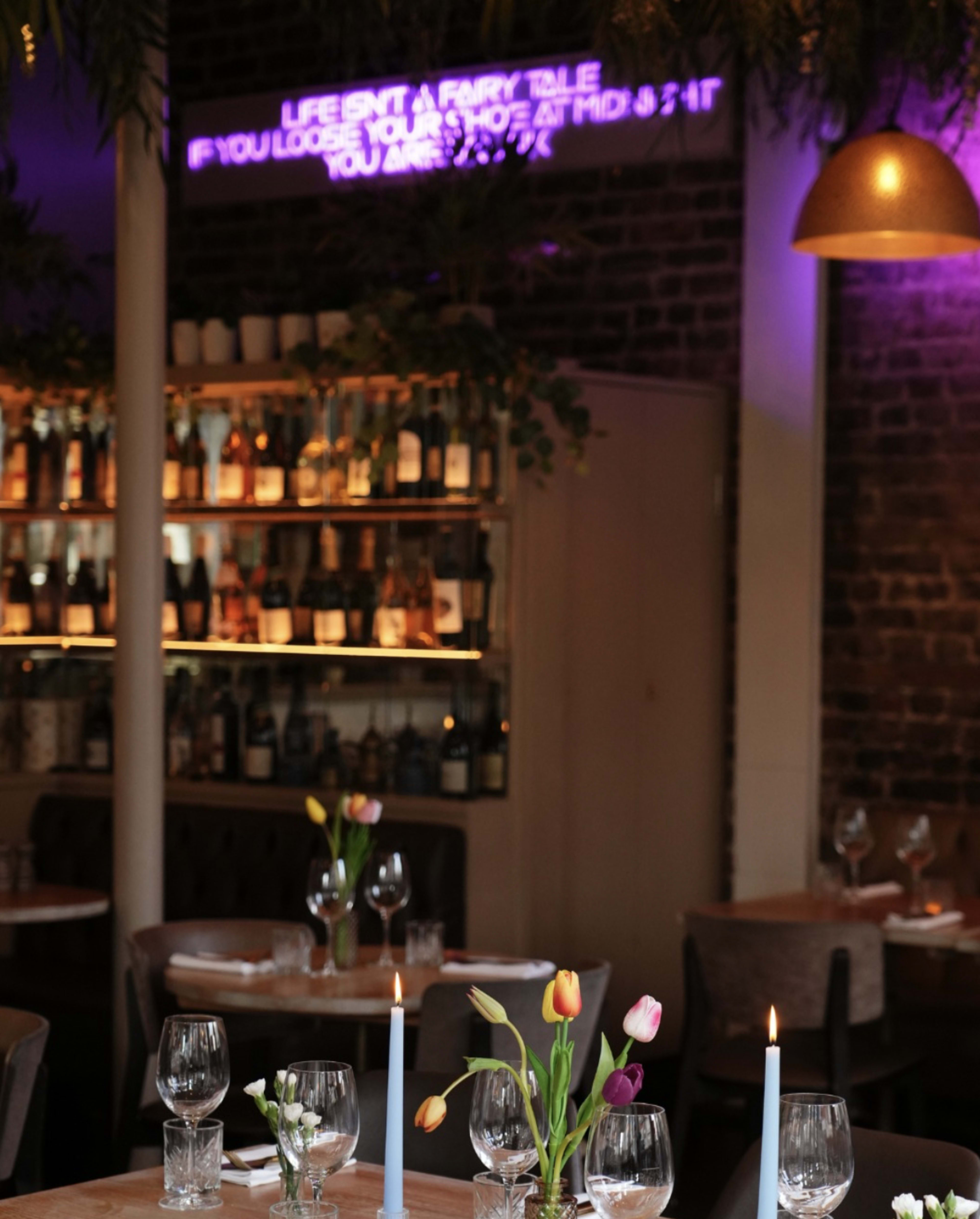The image shows a cozy restaurant interior with a neon sign, shelves filled with bottles, and tables set with flowers and candles.
