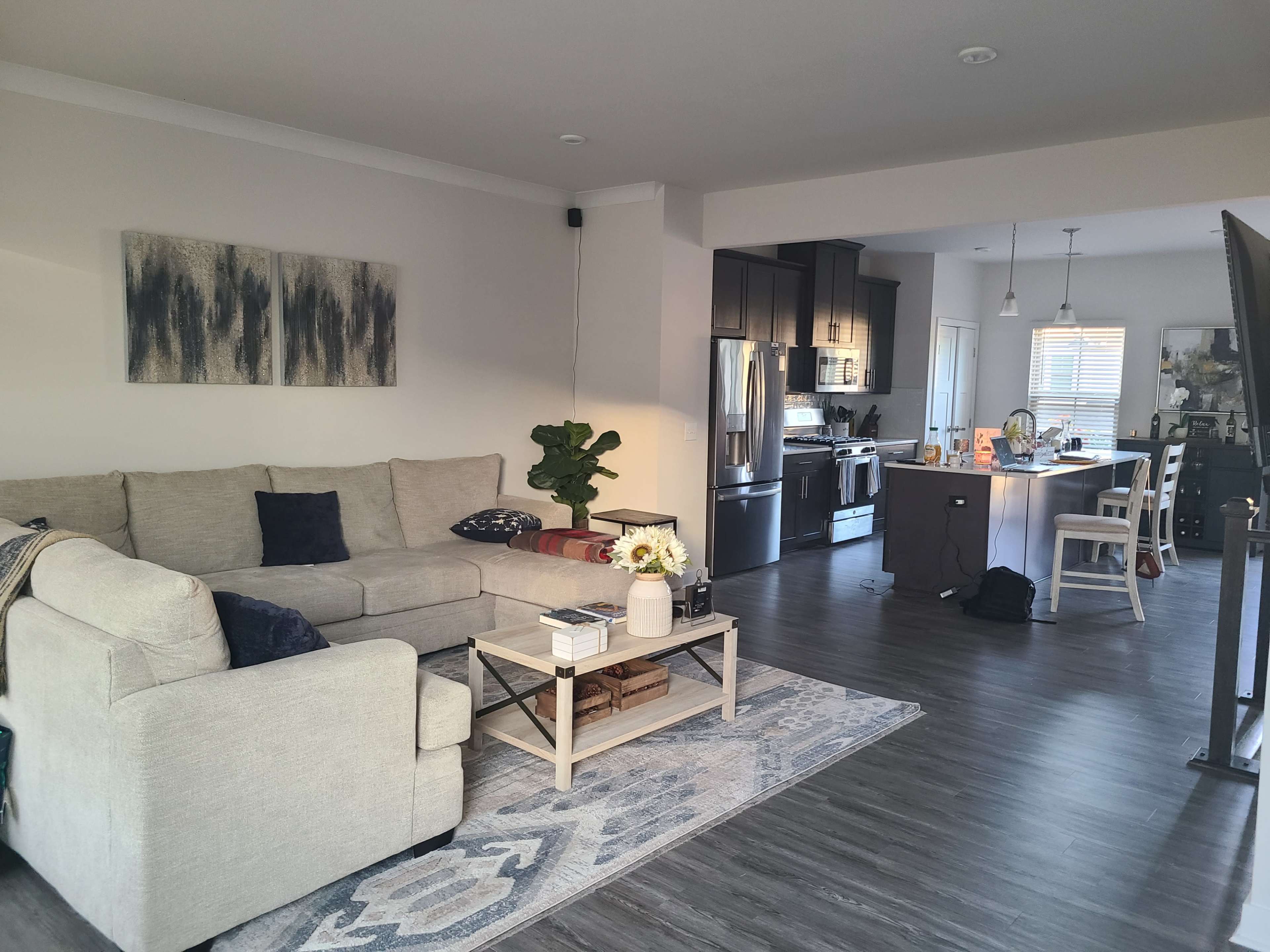 A living room with a light-colored sectional sofa, a coffee table, and an adjacent kitchen area featuring dark cabinetry and stainless steel appliances.