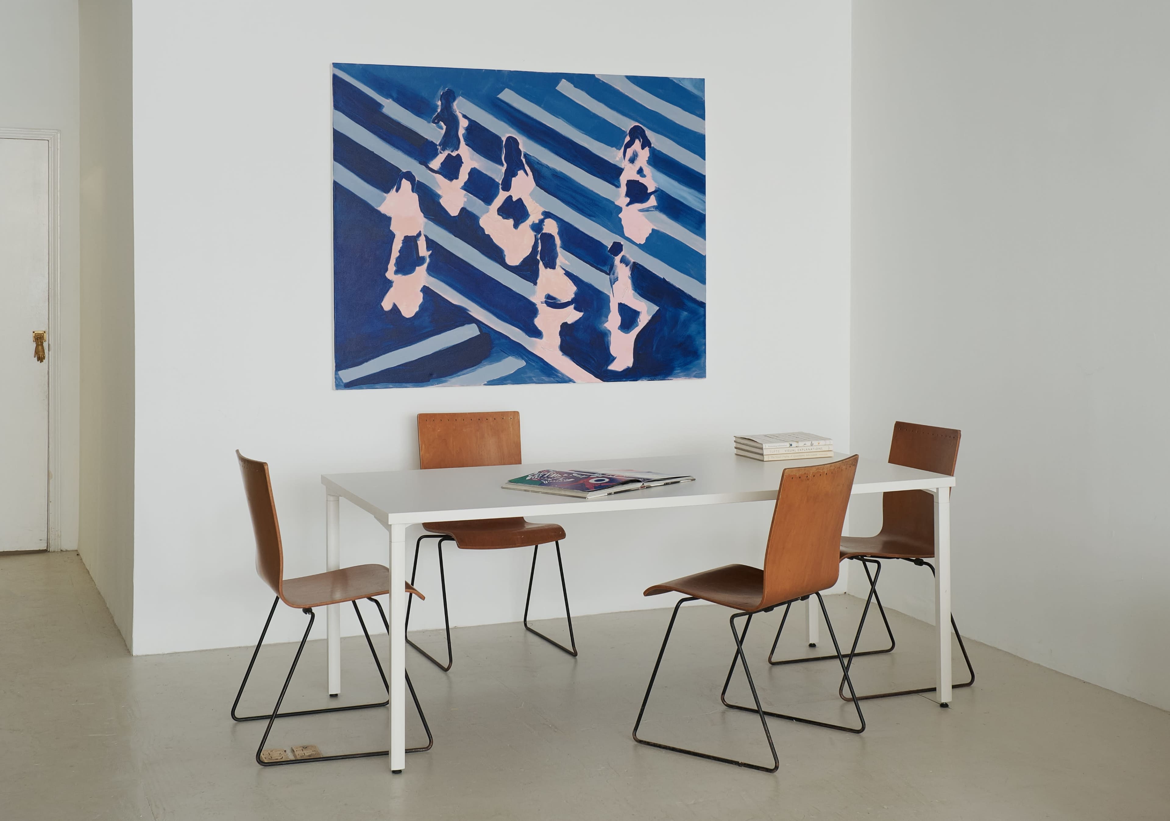 A minimalist dining area features a rectangular white table surrounded by four wooden chairs and a large blue and pink abstract painting on the wall.