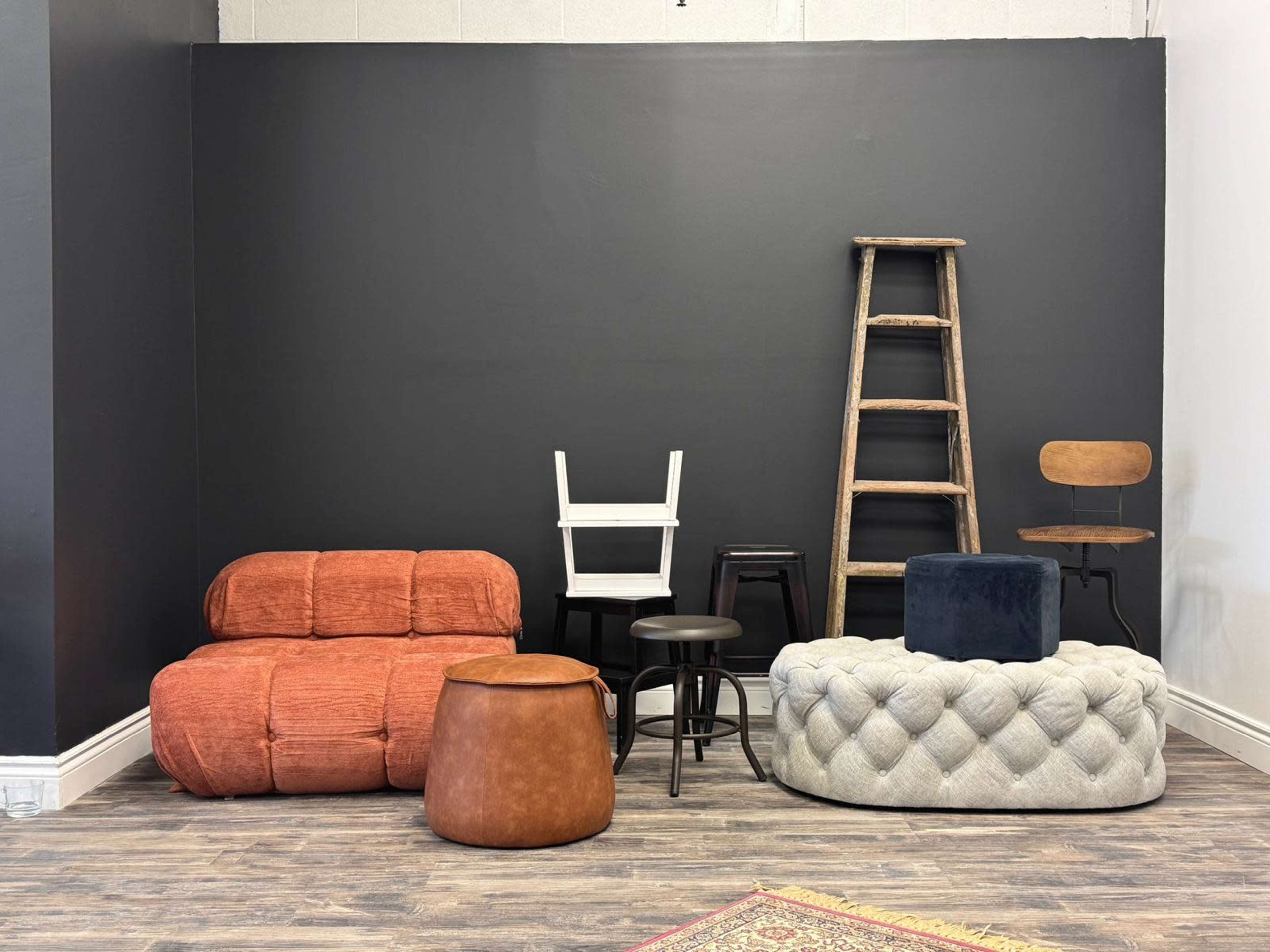 The image features a variety of furniture, including an orange upholstered sofa, several stools, a wooden ladder, and a round ottoman, arranged against a black wall.