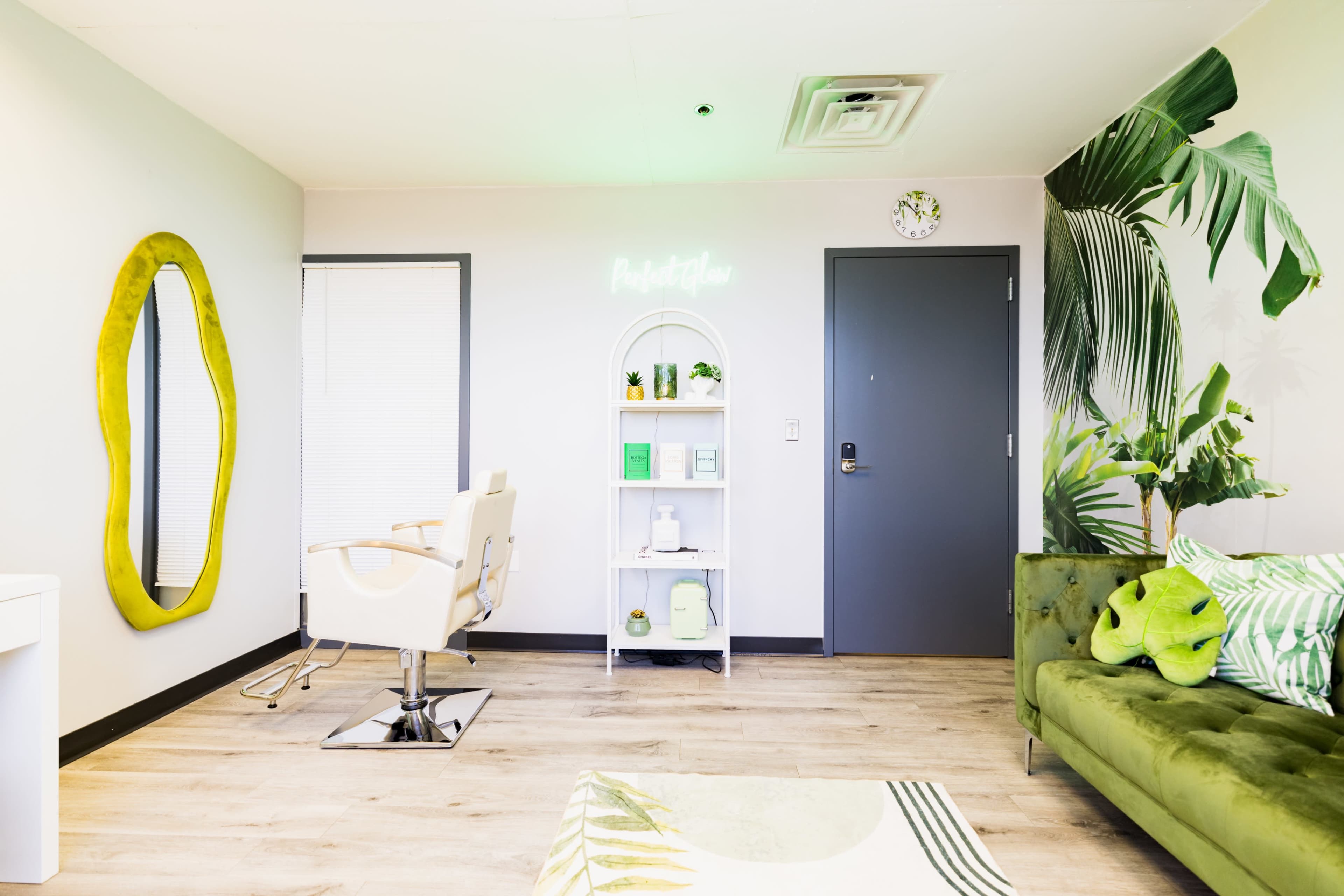 The image shows a bright, modern salon interior featuring a styling chair, a mirror, a green couch, and decorative plants on the walls.