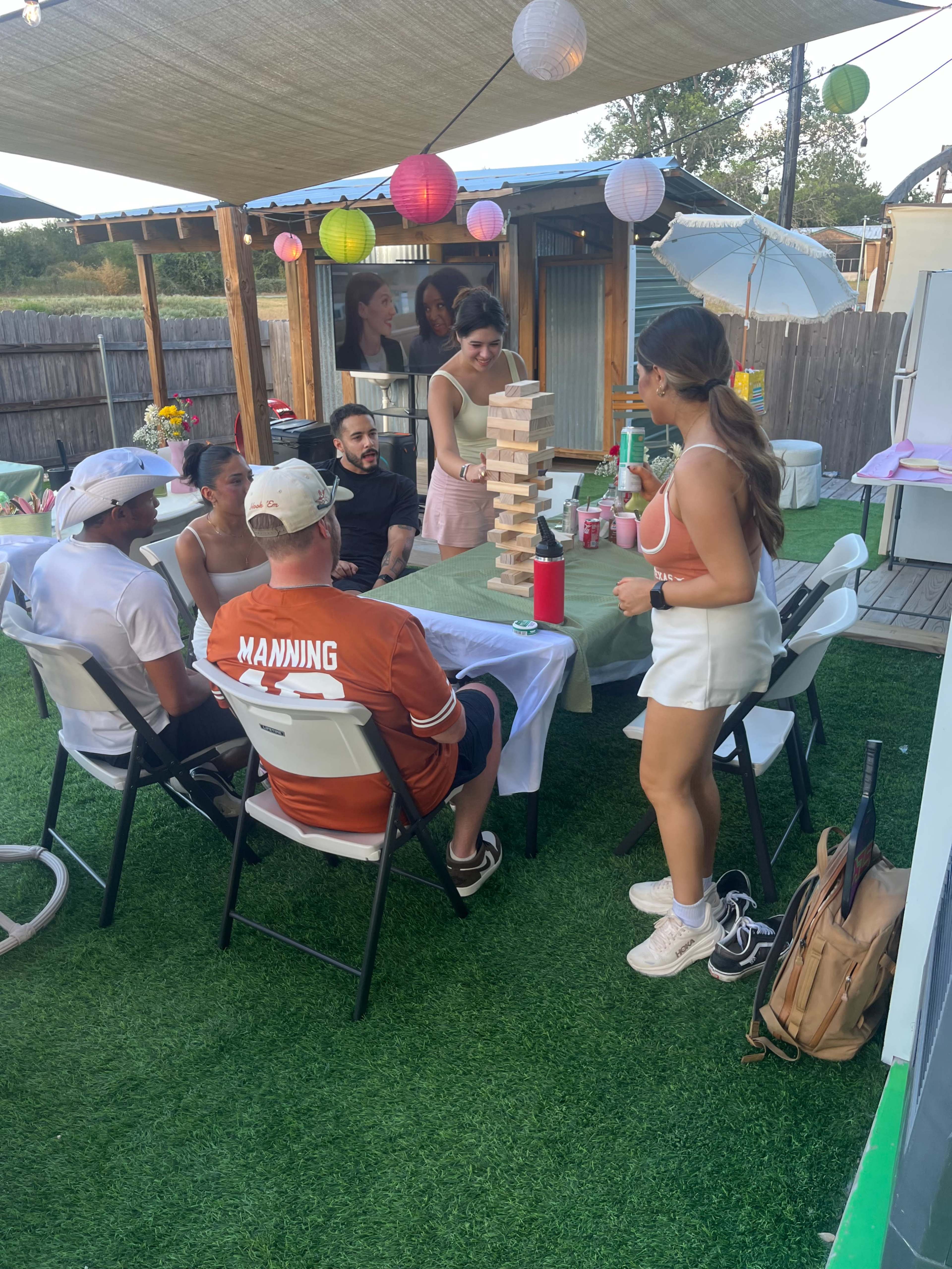 A group of people is gathered around a table outdoors, playing a game of Jenga with colorful lanterns hanging above.