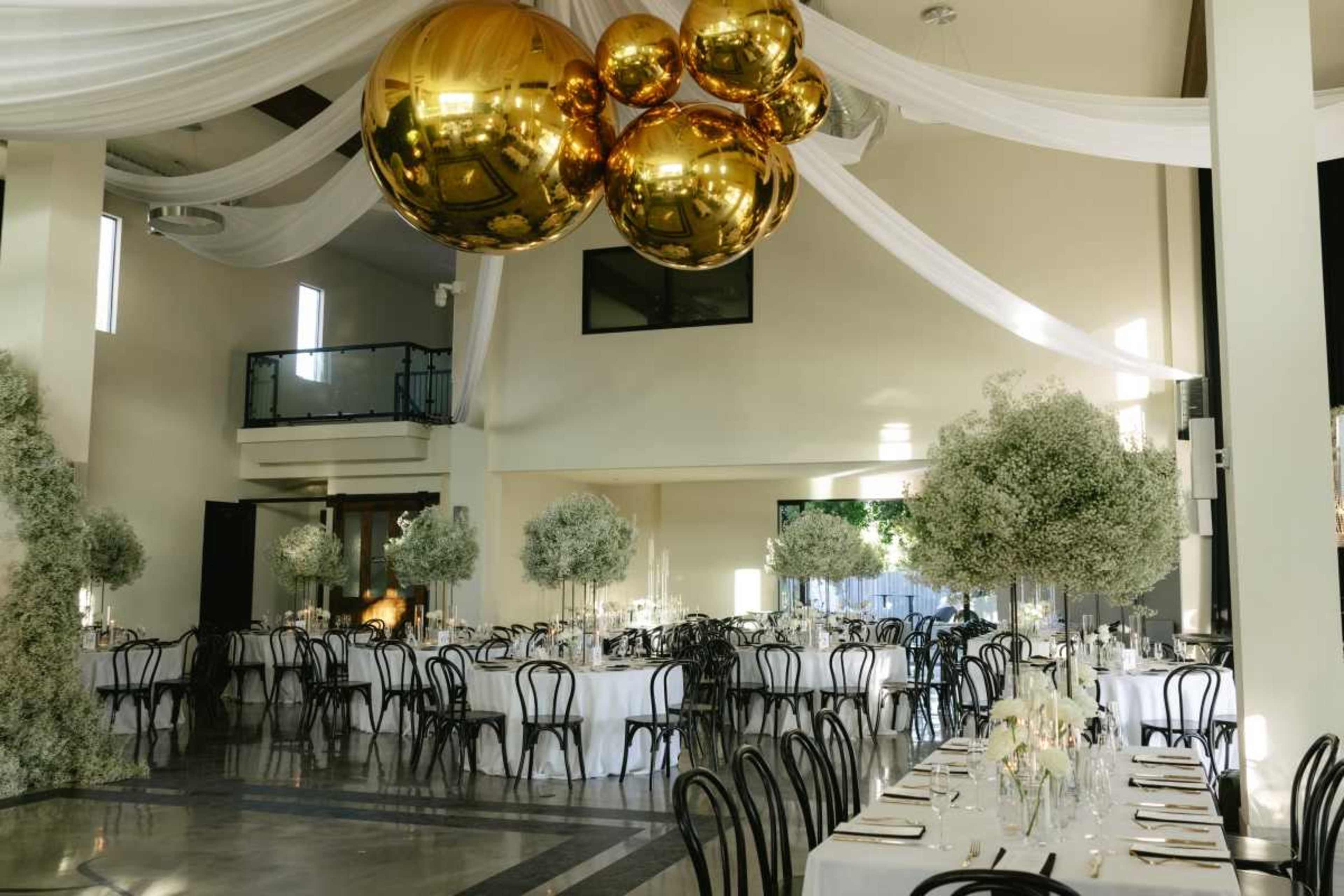 A spacious dining area features long tables set with white tablecloths and elegant centerpieces, surrounded by greenery and adorned with large golden balloons overhead.