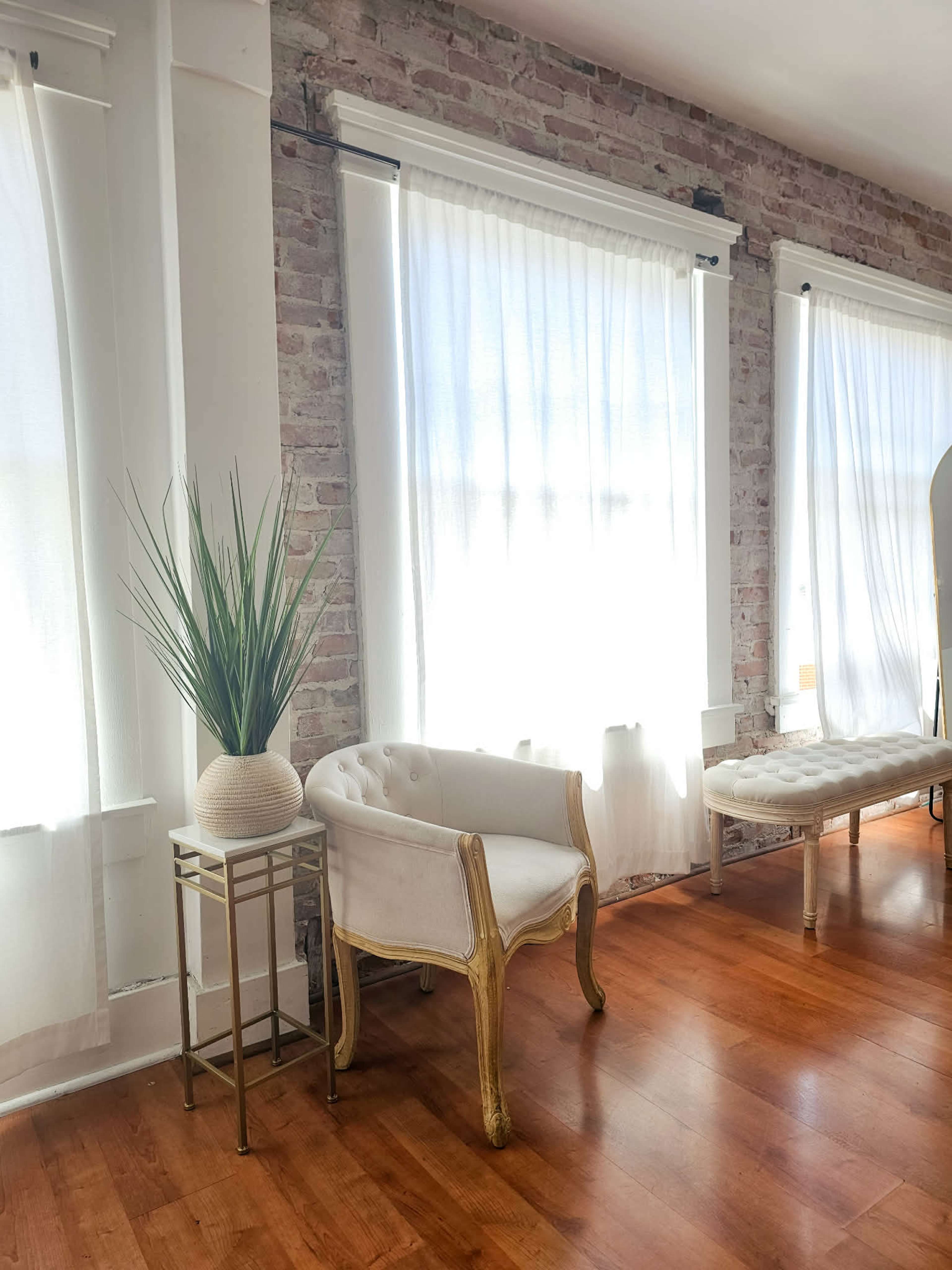 A bright room features large windows with sheer curtains, a white upholstered chair with gold accents, and a potted plant on a small metallic table.