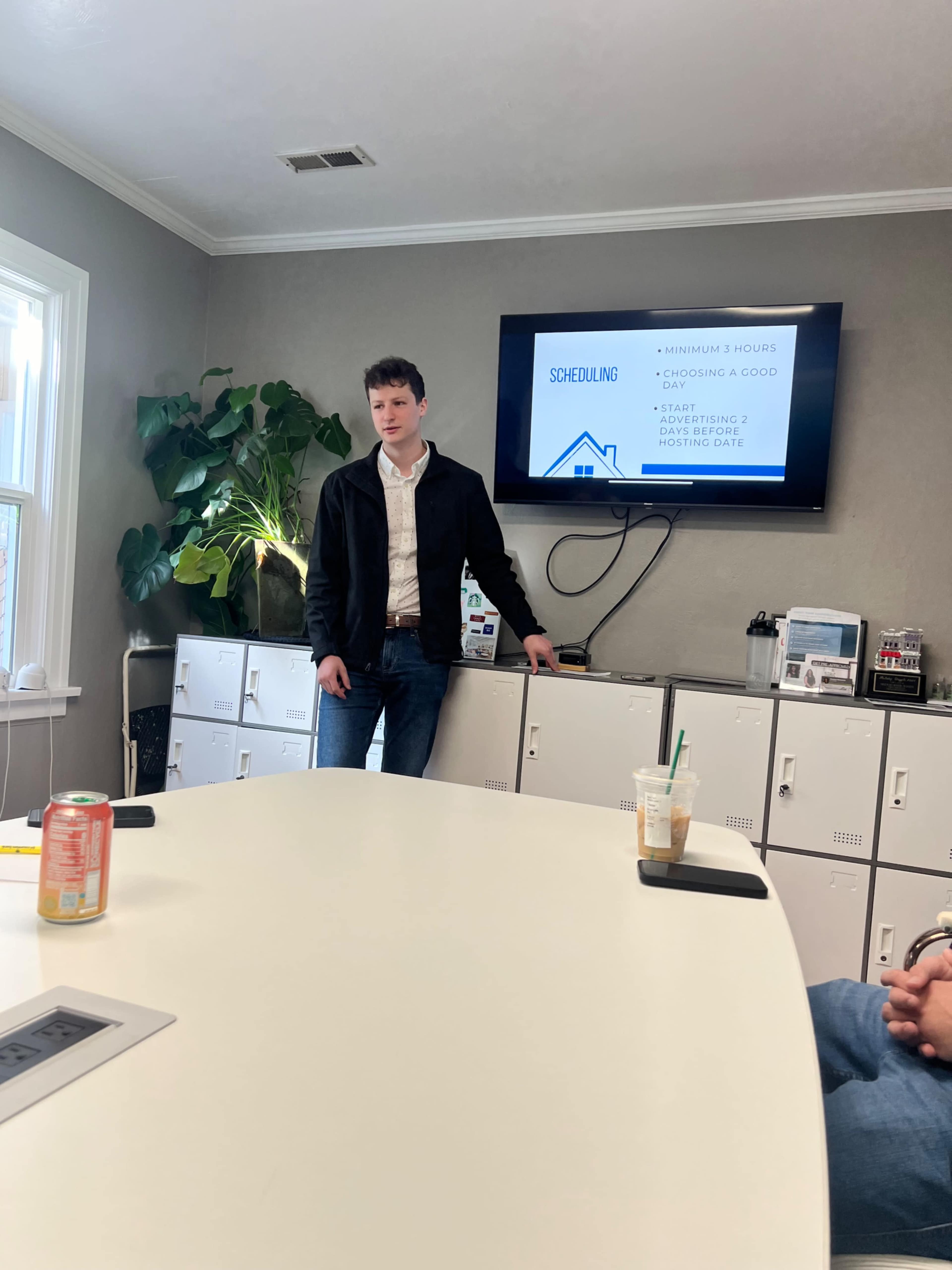 A young man stands in front of a presentation screen while speaking to a group in a conference room.