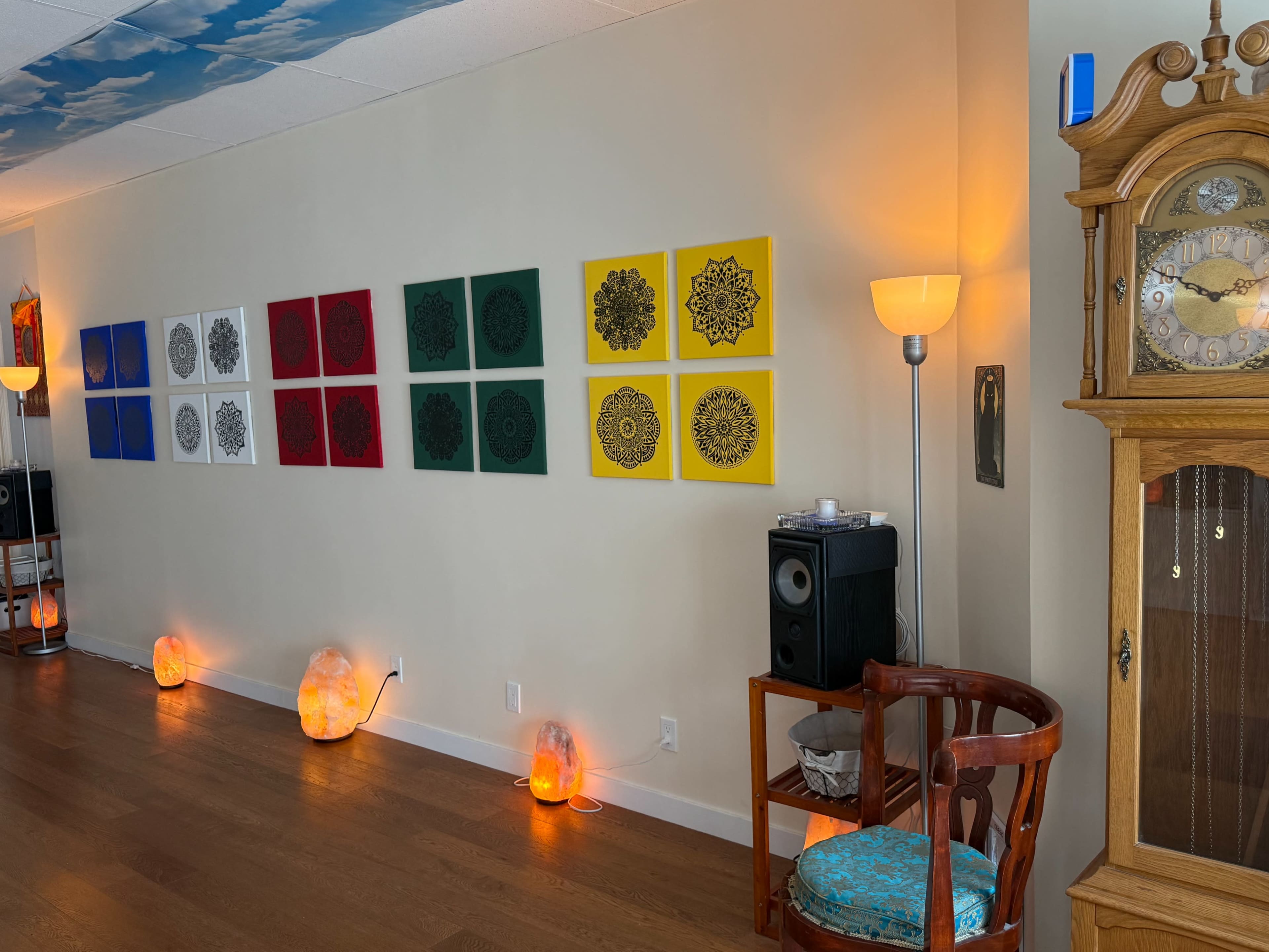 The image shows a brightly lit room with a wall displaying a series of patterned square canvases in various colors, along with a wooden clock and a speaker next to a chair, while salt lamps illuminate the floor.