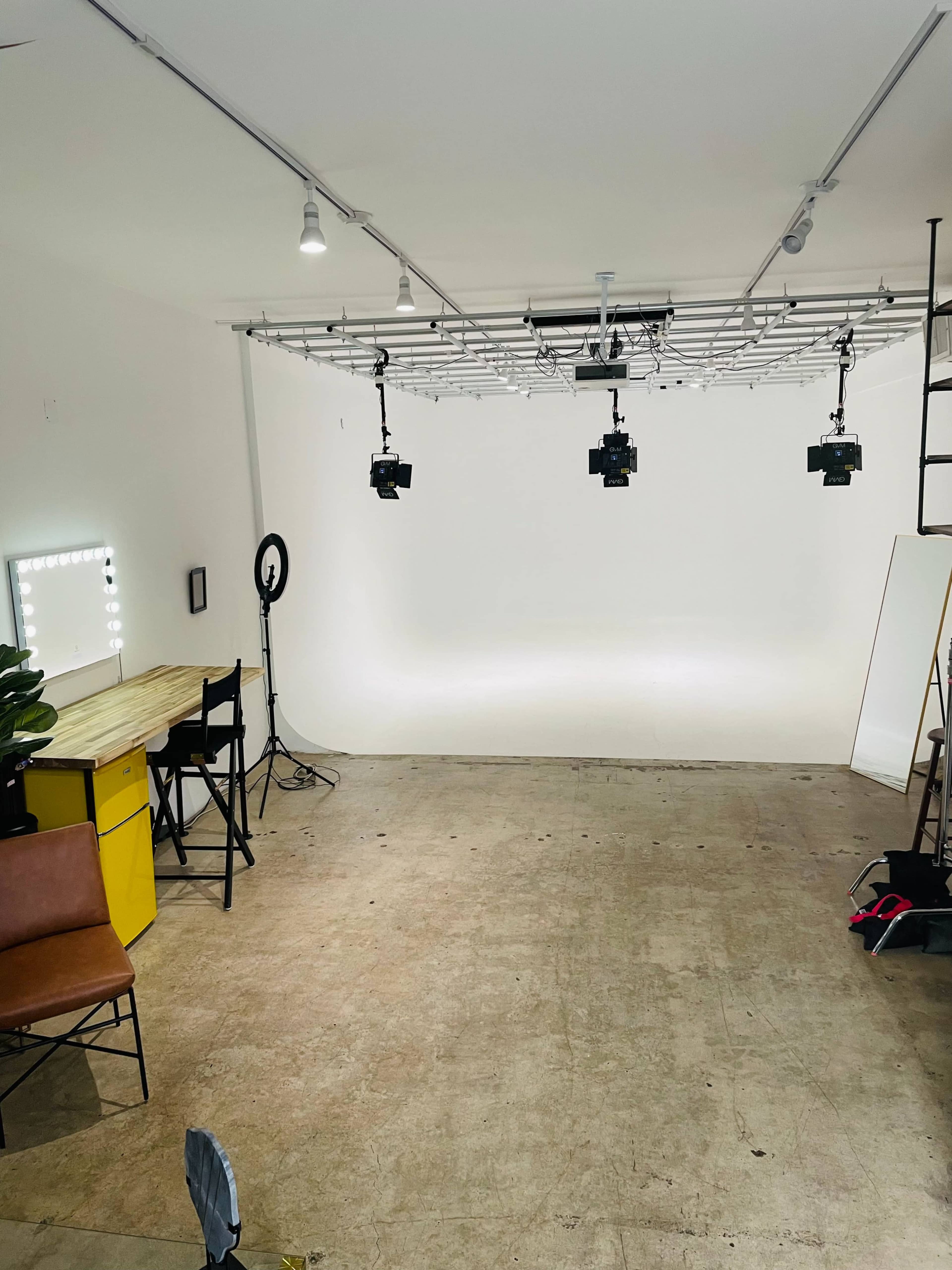 A minimalist studio space with a white backdrop, multiple lighting setups, a desk, and a chair.