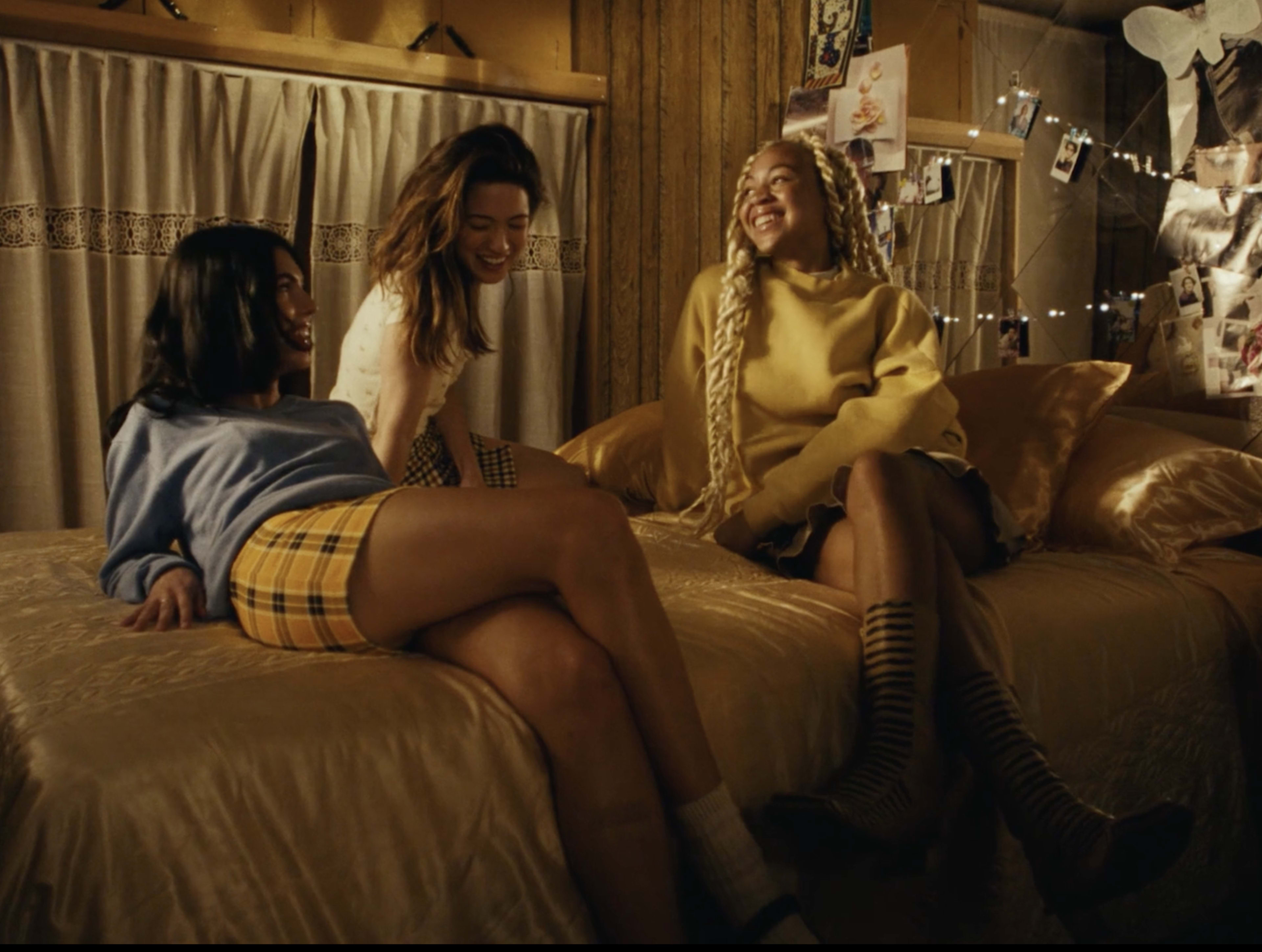 Three young women are sitting on a bed in a cozy room decorated with photos and lights, smiling and engaging in conversation.