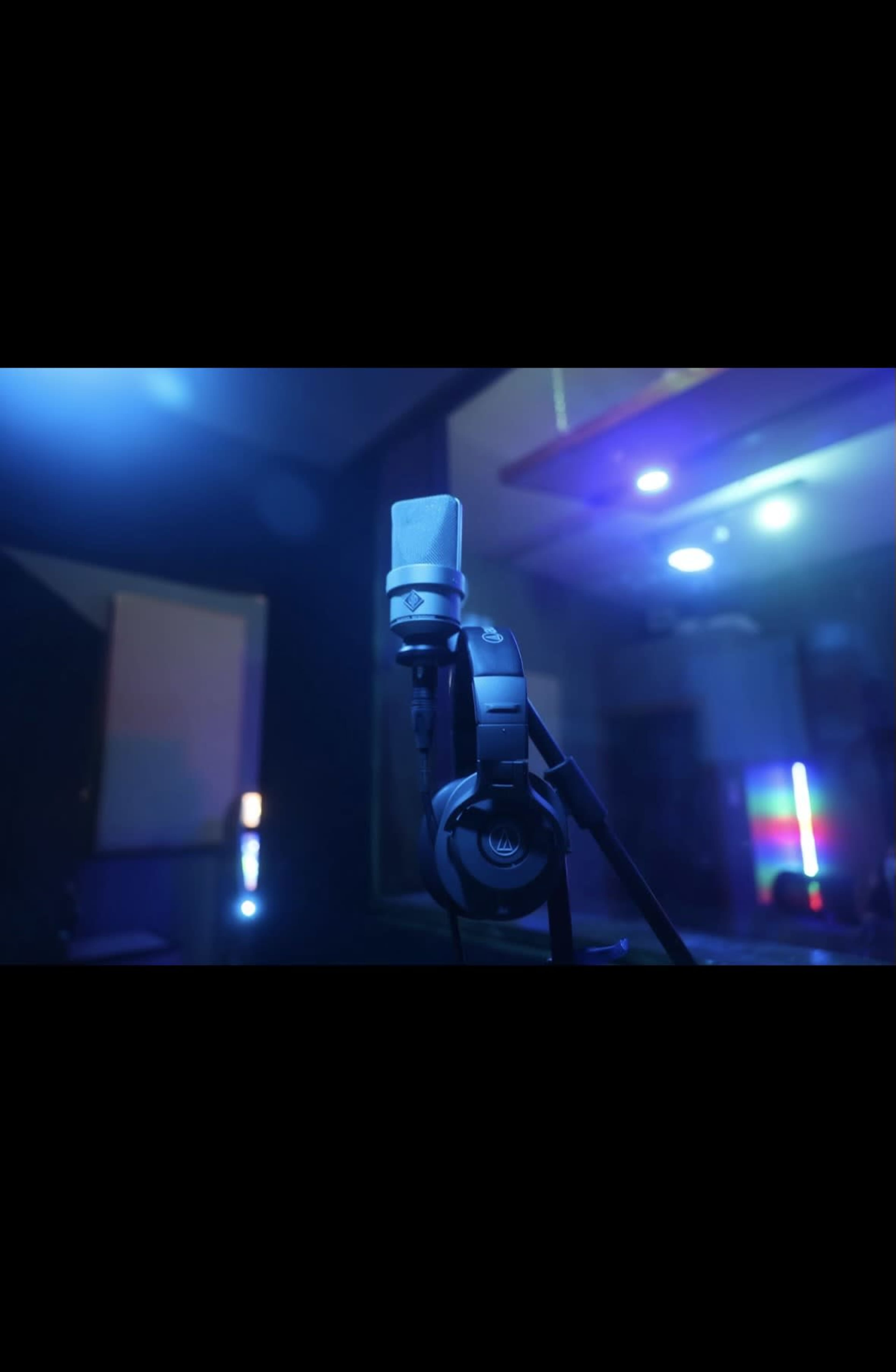A microphone with headphones attached stands on a stand in a dimly lit recording studio, illuminated by colored lights.