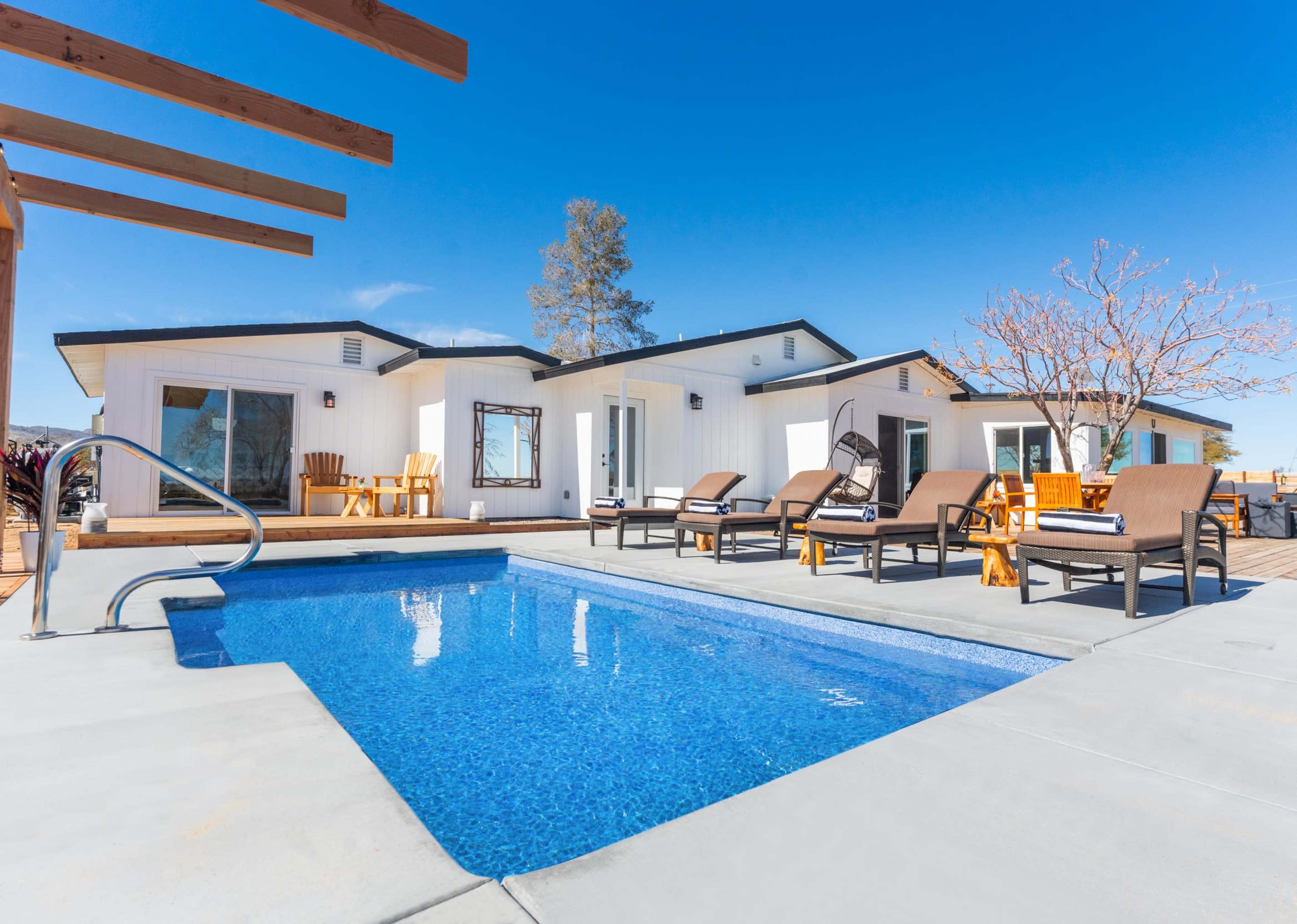 The image shows a modern outdoor area featuring a small pool surrounded by lounge chairs and two white houses under a clear blue sky.