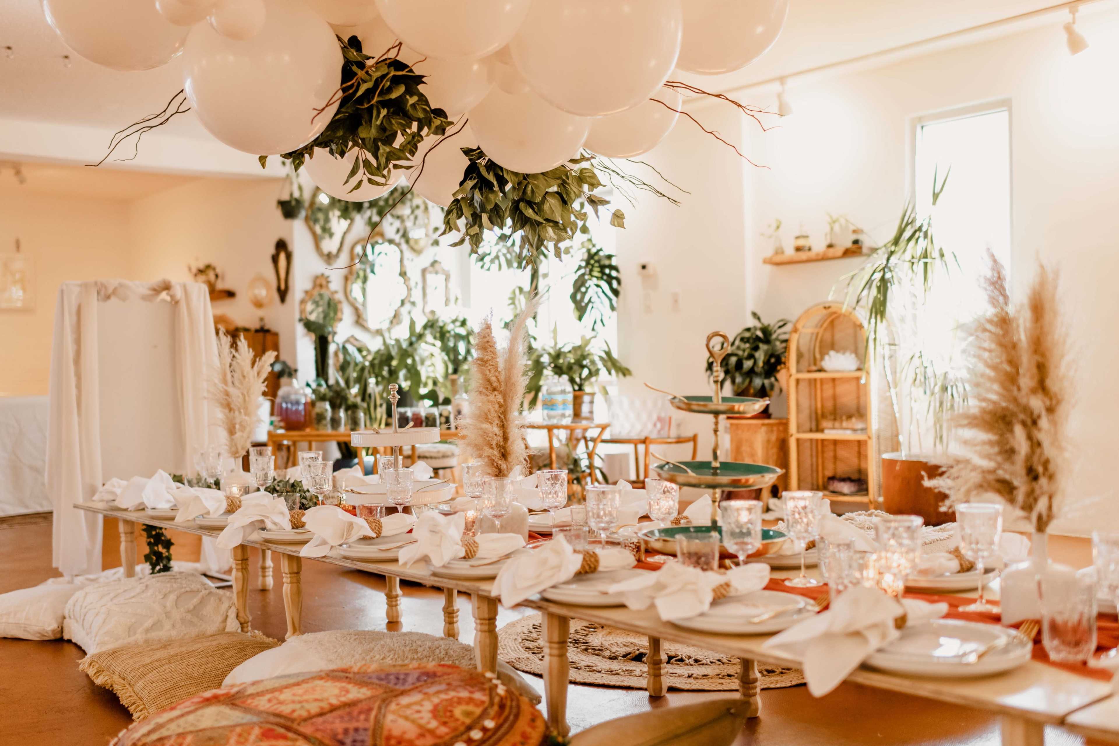 The image shows a beautifully decorated indoor dining setup featuring a long table with floral arrangements, elegant tableware, and a canopy of white balloons overhead.