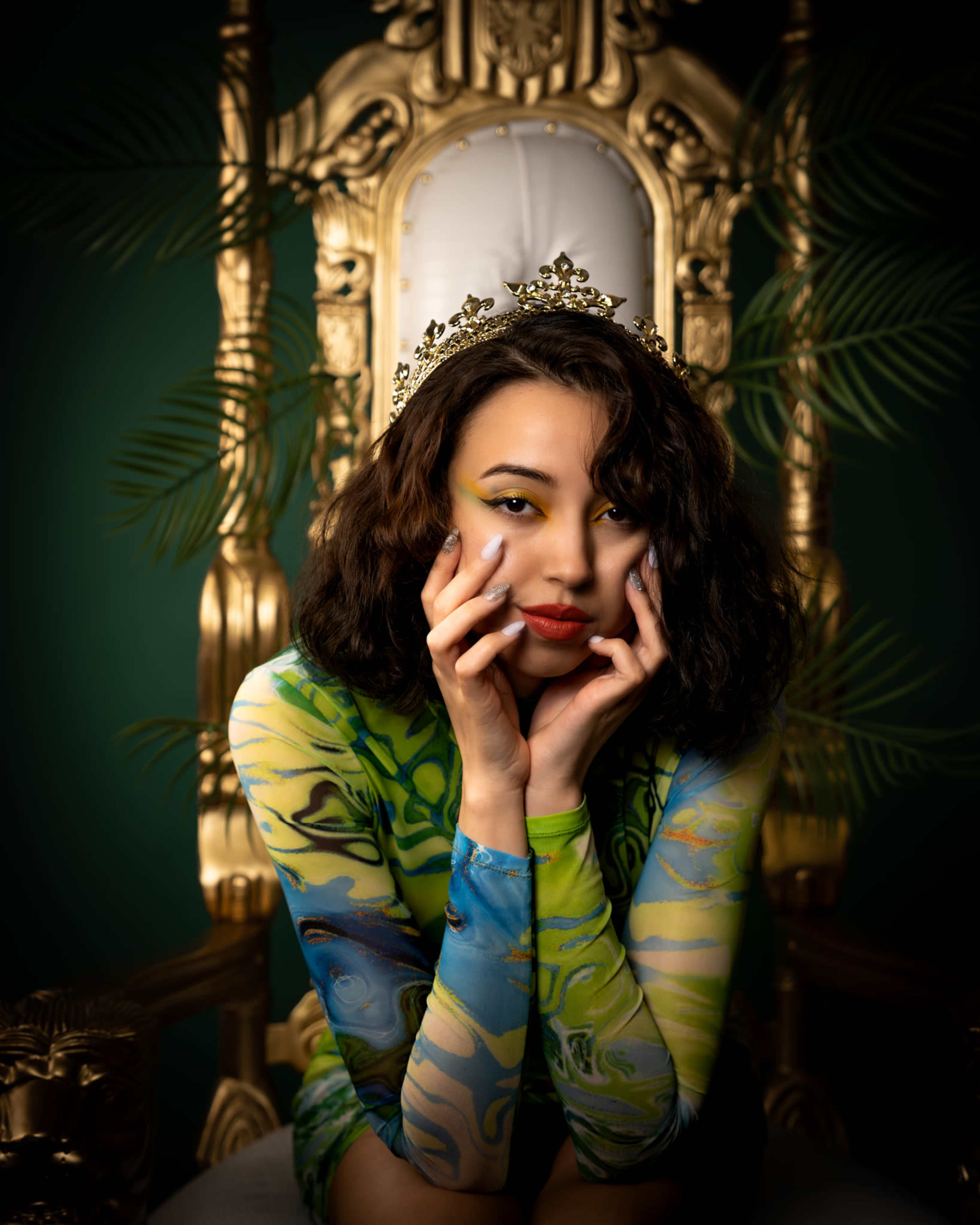 A person in a patterned outfit sits on a gold throne, holding their face with both hands and wearing a tiara.