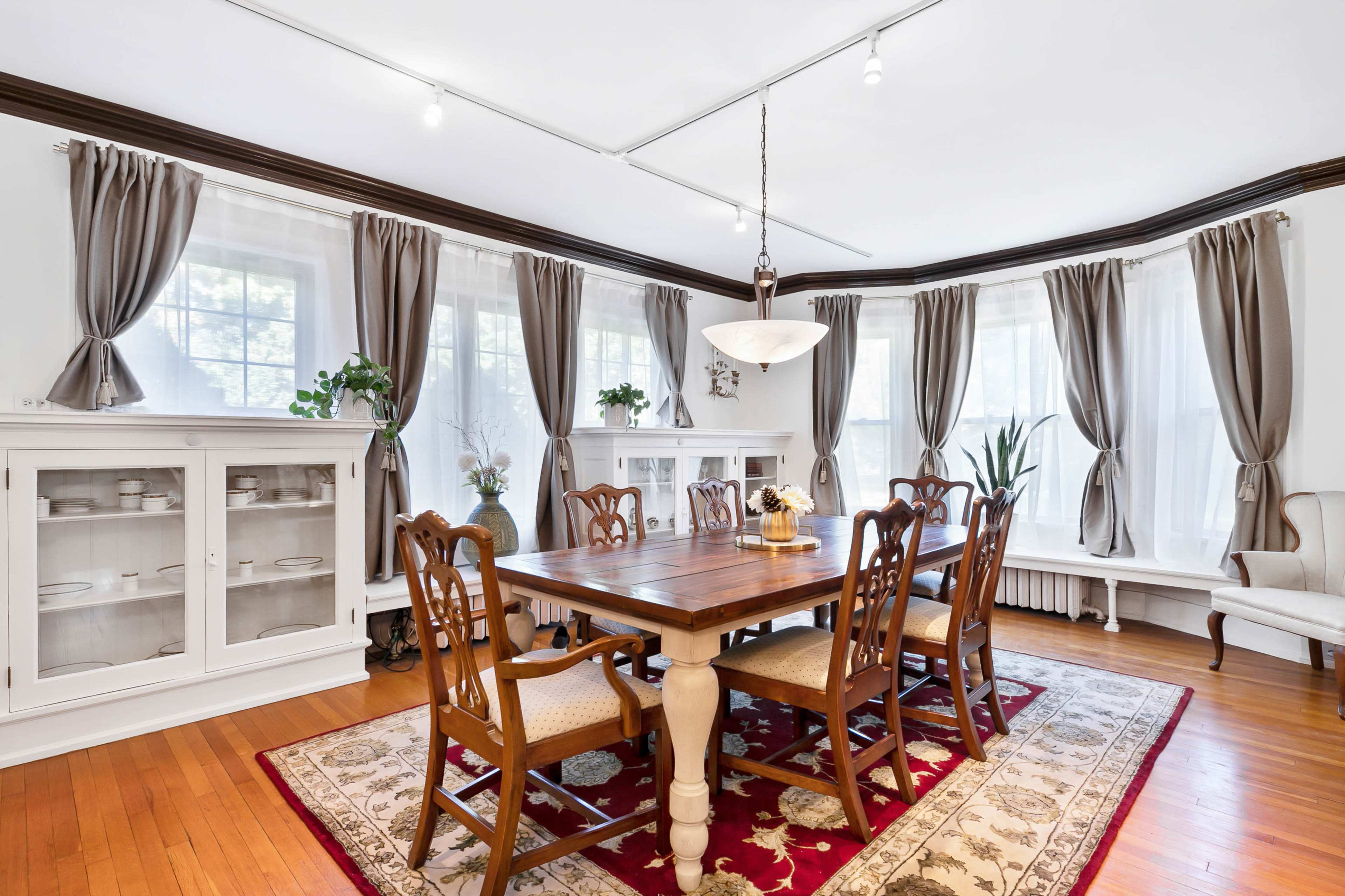 A wooden dining table surrounded by chairs is set in a well-lit room featuring bay windows, white cabinetry, and draped curtains.
