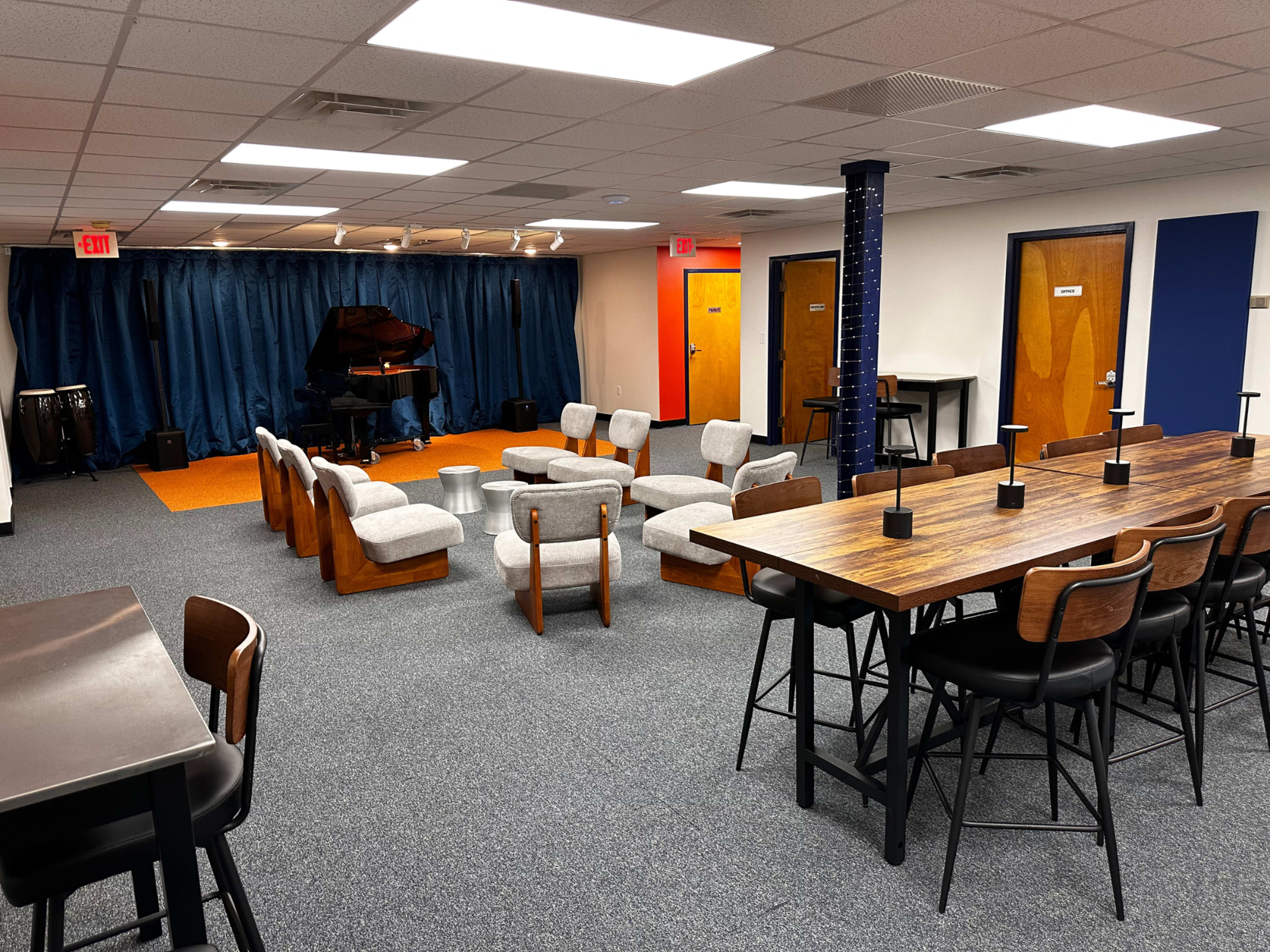 The image shows a spacious room with a seating area arranged in a semi-circle, a long dining table with chairs, and a grand piano on a stage behind a blue curtain.