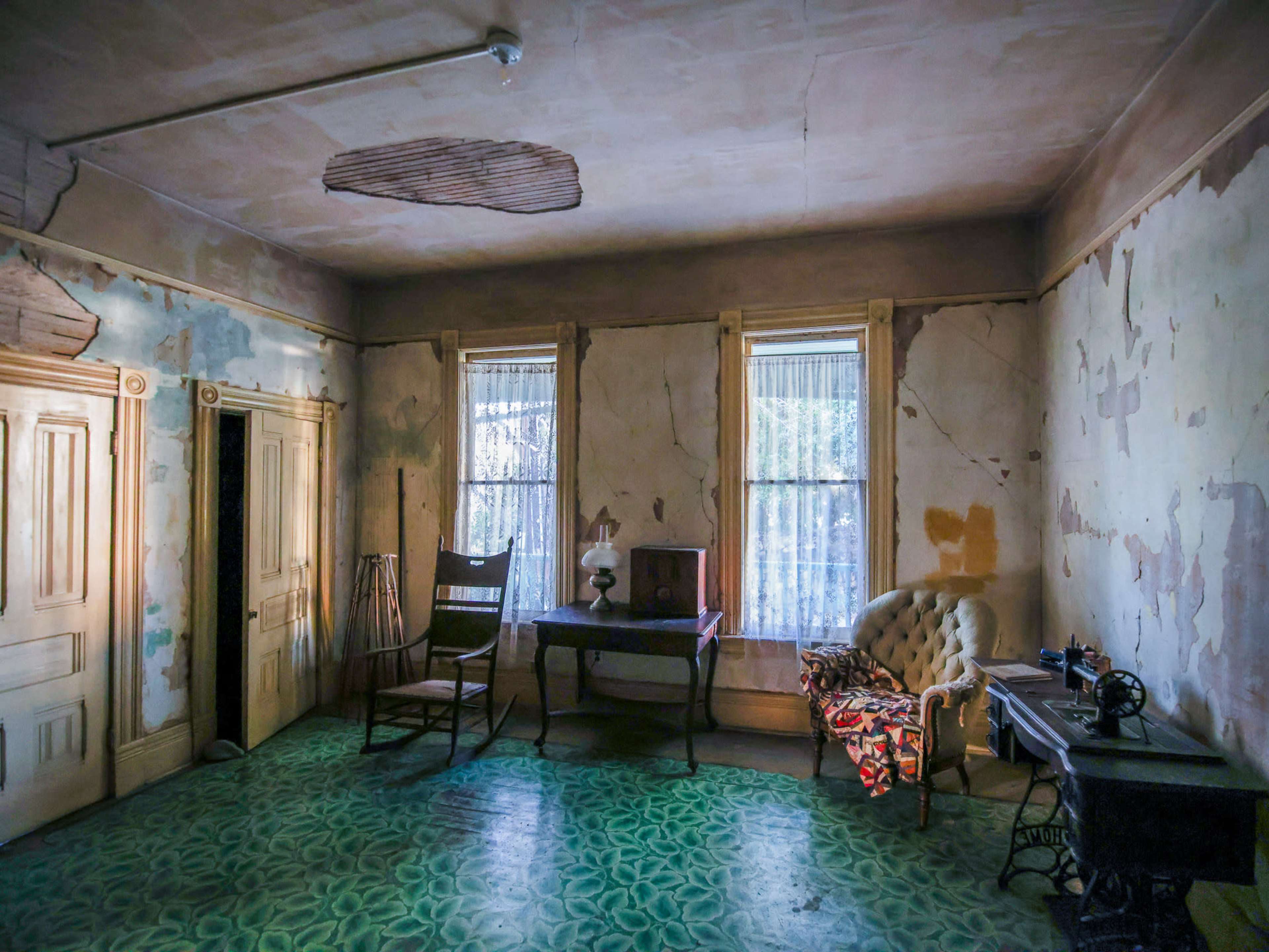 An empty room with peeling walls, two windows, a wooden desk, a chair, and an old sewing machine.
