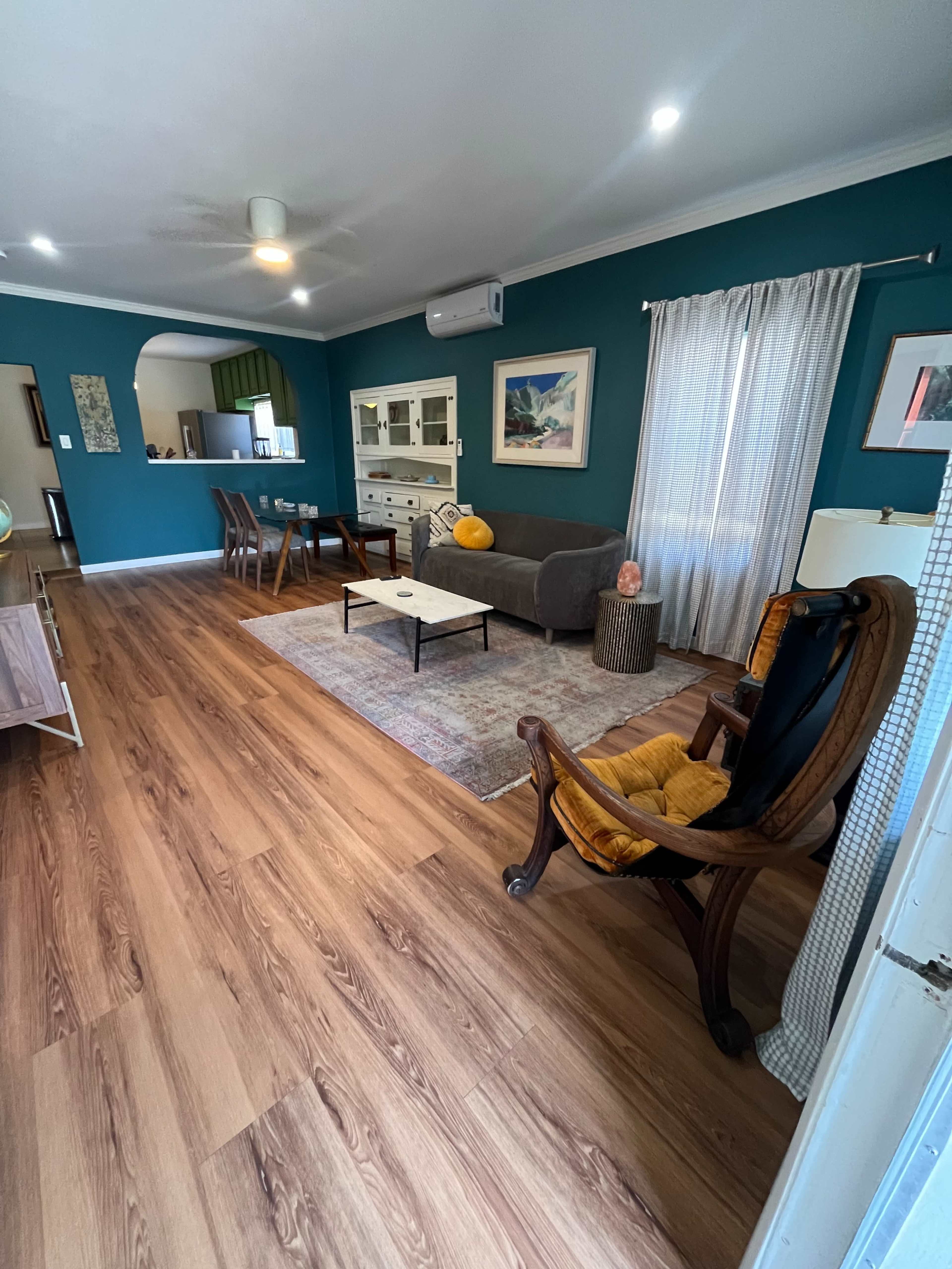 The image shows a living room with a gray sofa, wooden chairs, a decorative rug, and teal walls, featuring a dining area in the background.