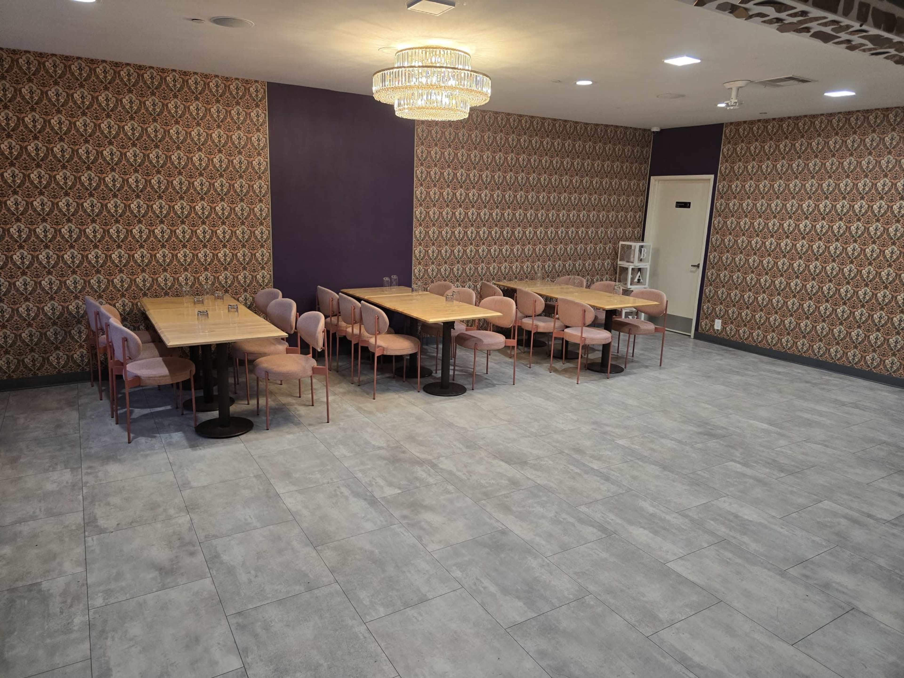 The image shows a spacious, brightly lit dining area with multiple tables and chairs arranged in two parallel rows, set against a patterned wallpaper backdrop.