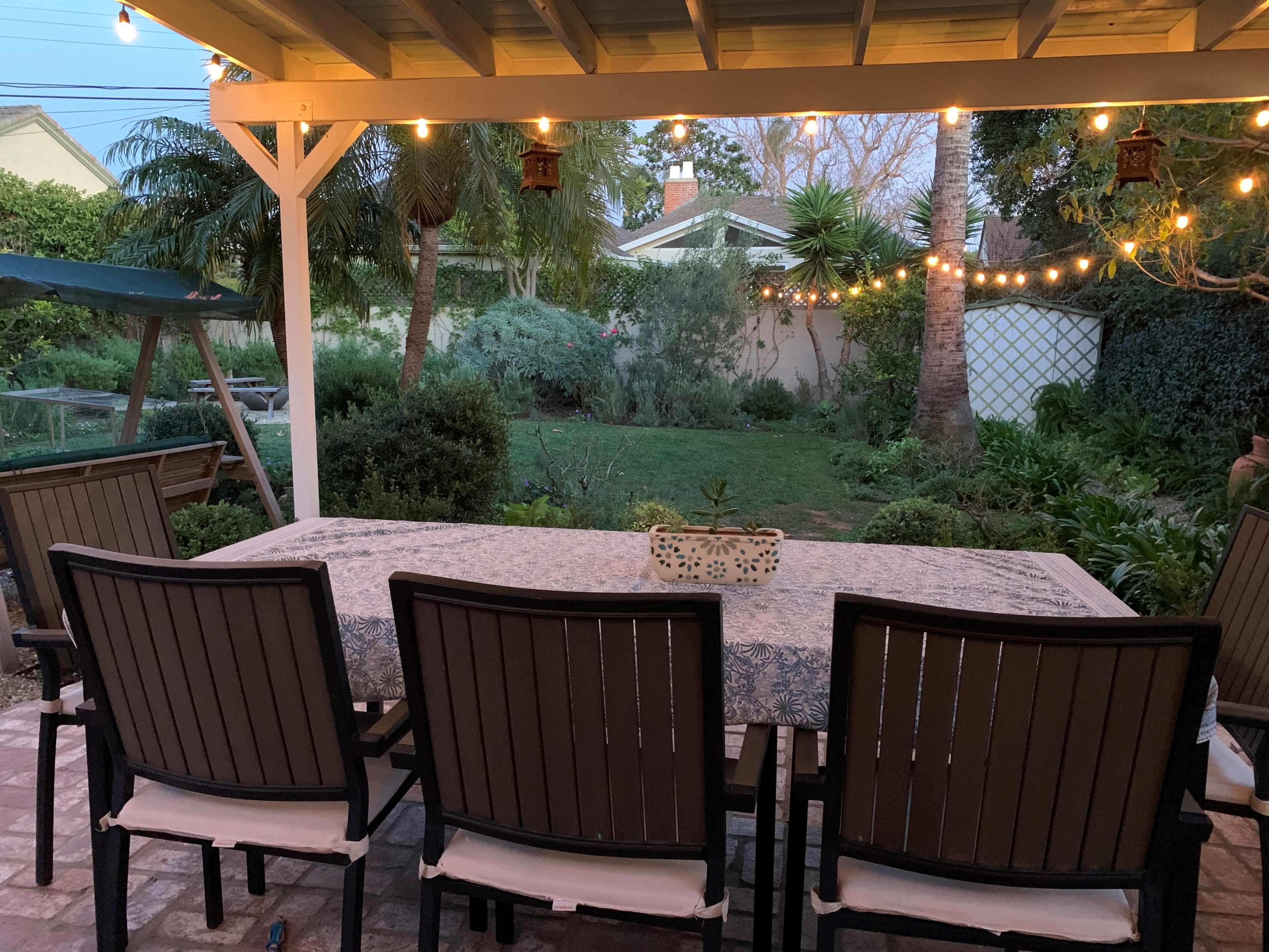 A backyard patio features a long dining table surrounded by chairs, with string lights overhead illuminating the lush garden in the background.