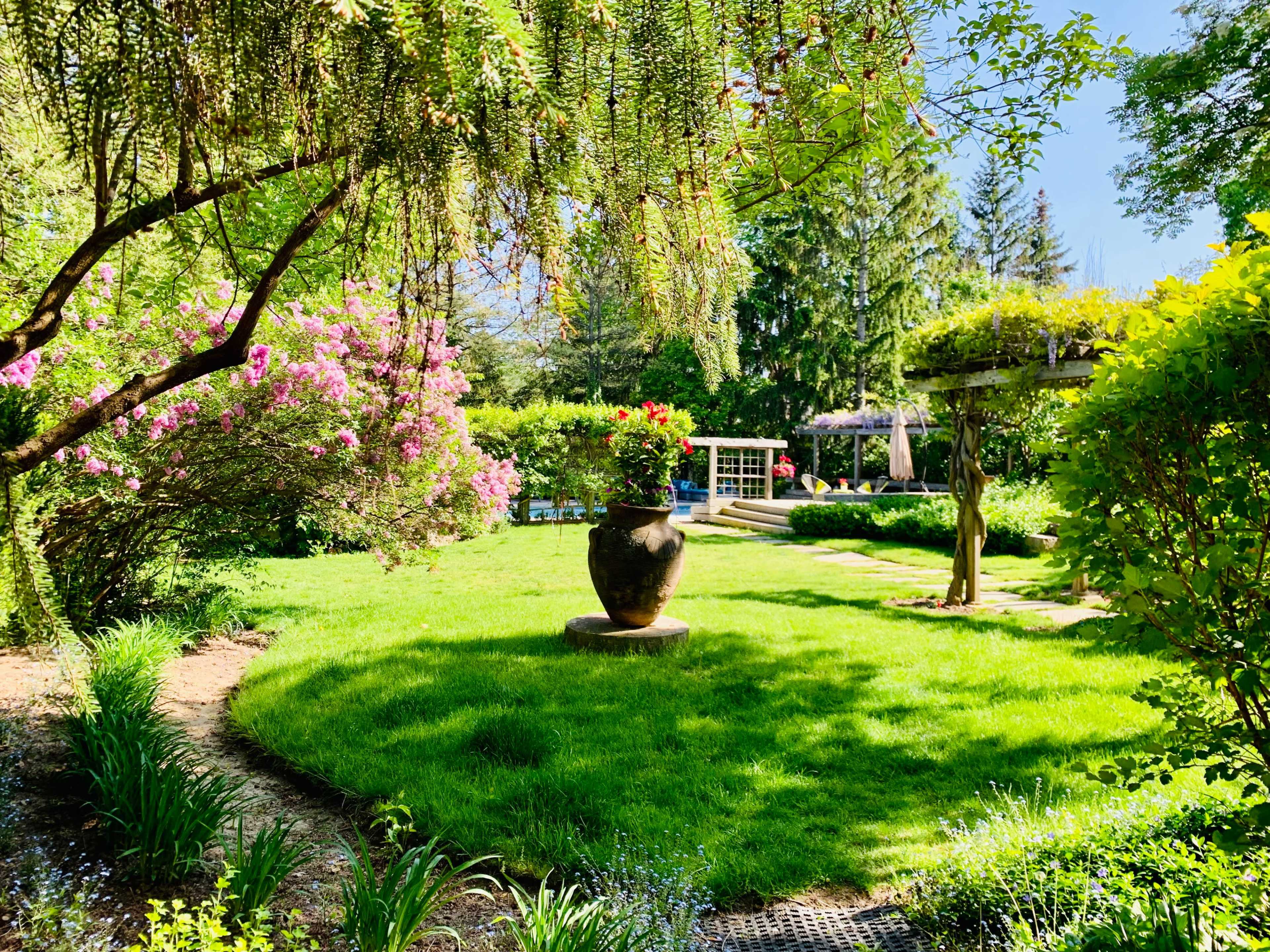 A landscaped garden features a circular lawn surrounded by flowering shrubs and a large decorative pot at the center.