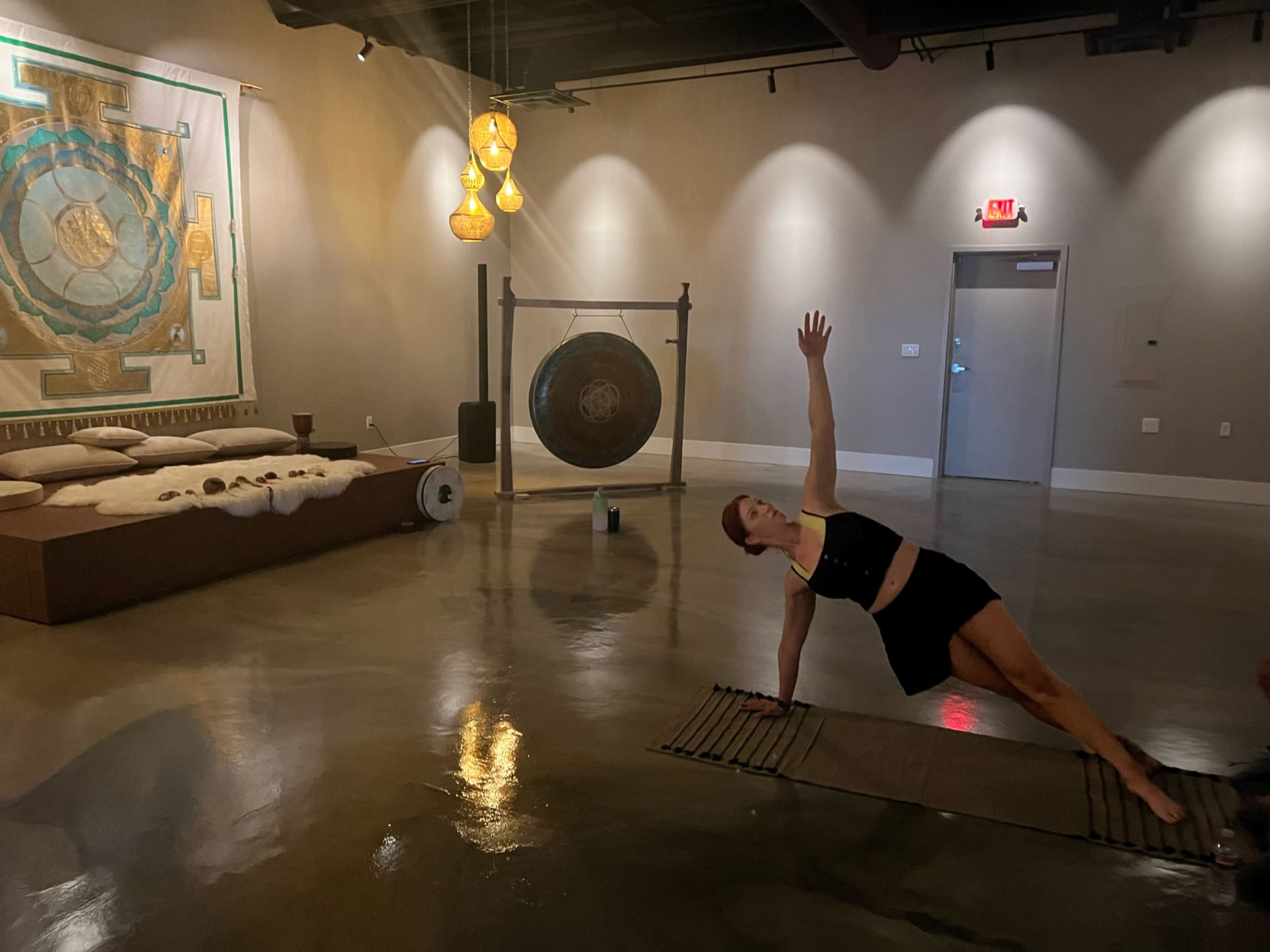 A woman performs a side plank exercise on a mat in a spacious, well-lit room with a large gong and decorative wall art.