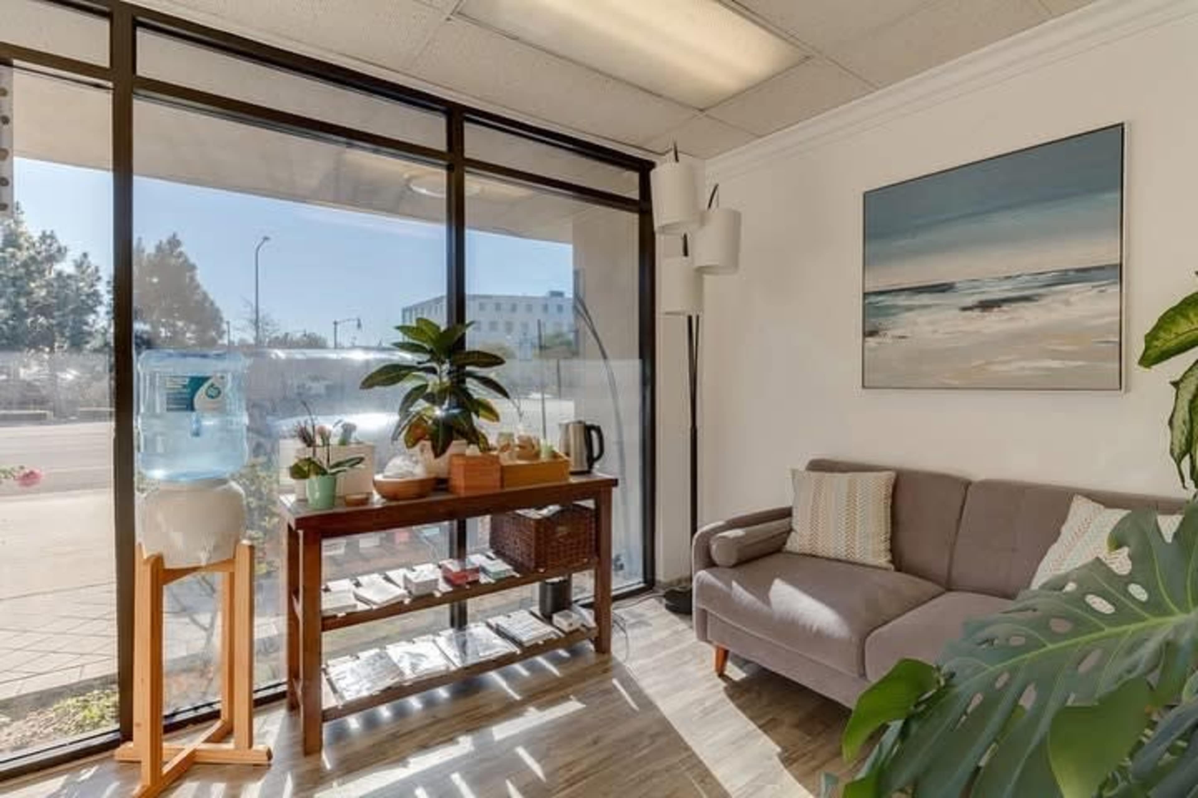 The image shows a well-lit waiting area with a gray couch, a wooden table featuring plants and a water cooler, and large windows providing a view of the outside.